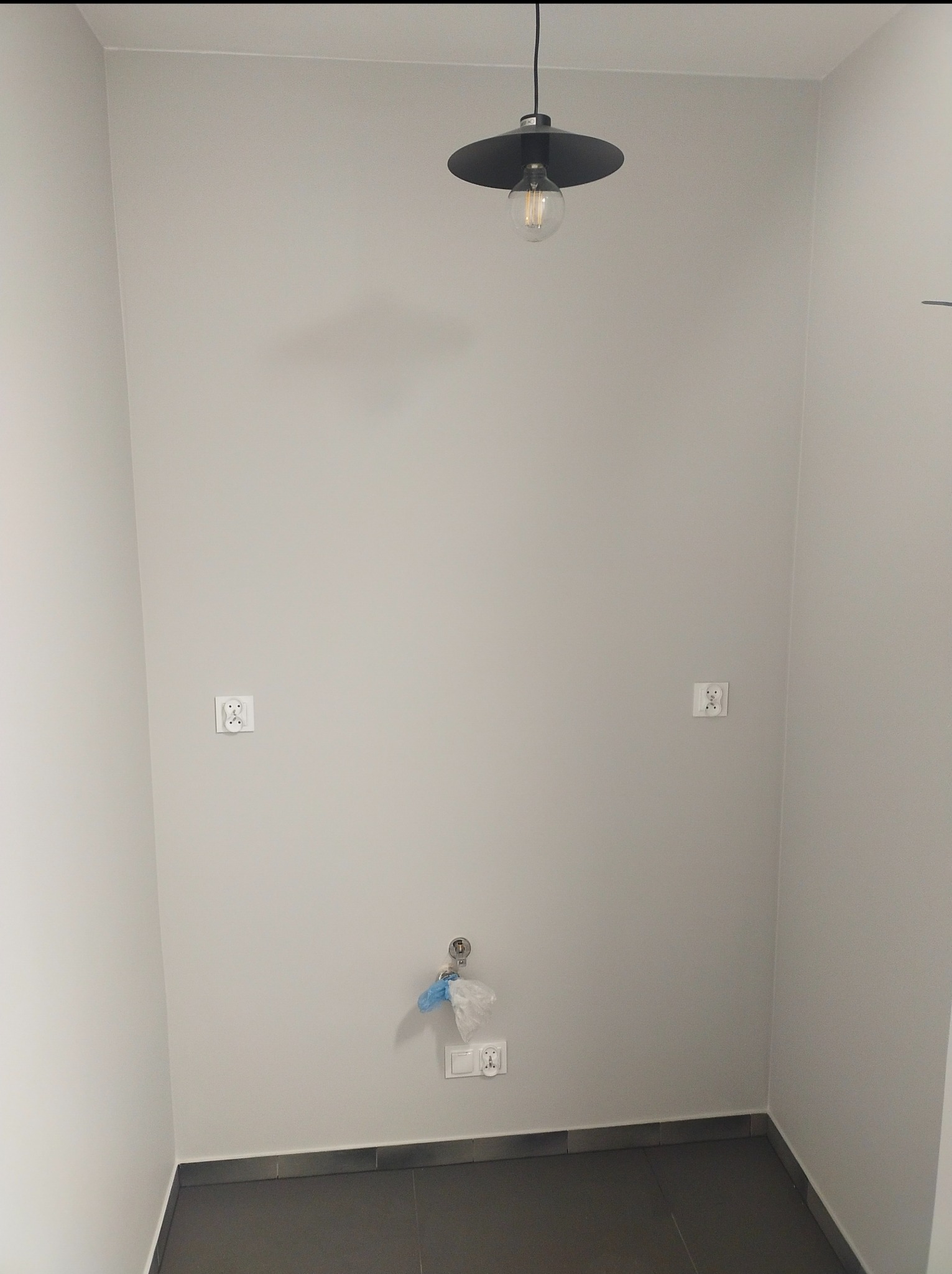 Pusta, wykafelkowana przestrzeń z szarą podłogą, przygotowana do montażu armatury łazienkowej, widoczne wystające przyłącza wody i gniazdka elektryczne, oświetlona lampą wiszącą z żarówką Edisona.
