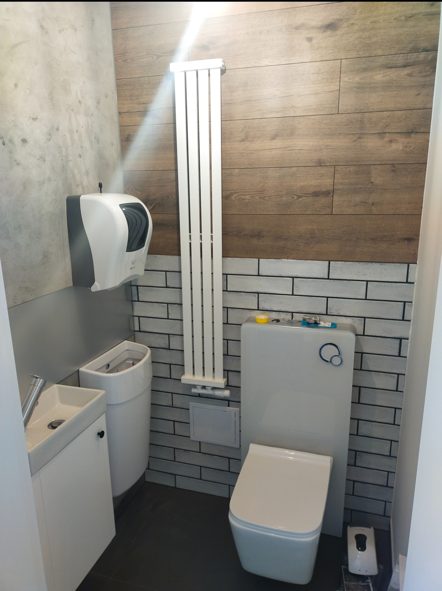 Nowoczesna toaleta z umywalką w małej łazience wykończonej płytkami imitującymi cegłę i drewno, grzejnik drabinkowy na ścianie.