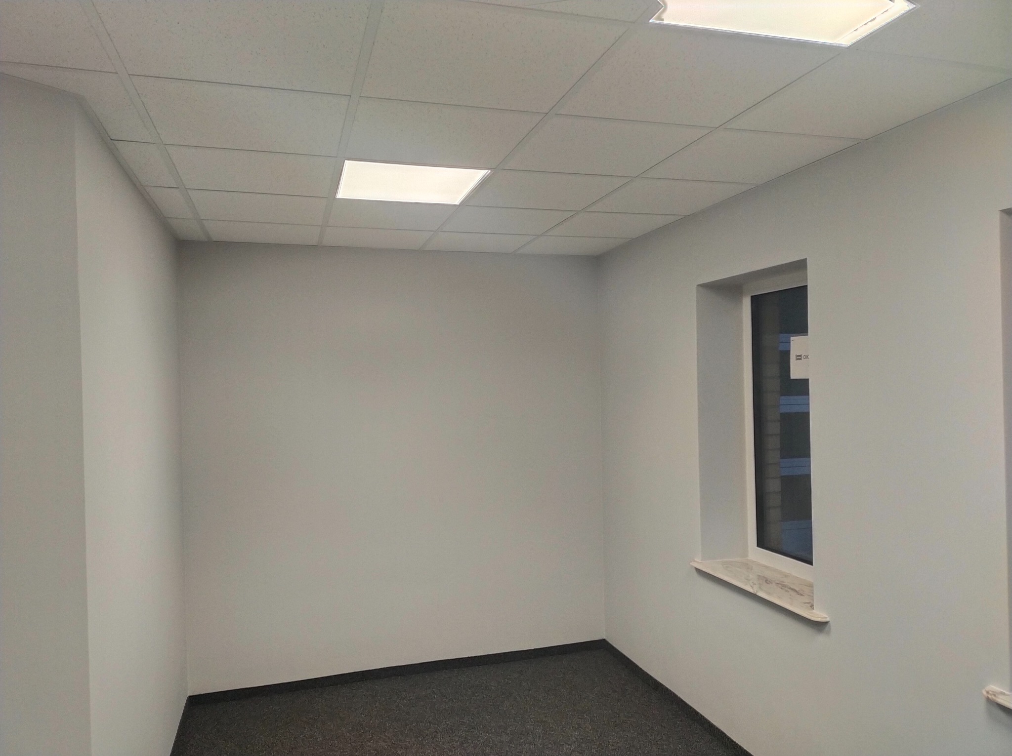 Pomalowane na szaro wnętrze pokoju z szarą wykładziną dywanową, białym oknem z marmurowym parapetem i kasetonowym sufitem z wbudowanym oświetleniem.