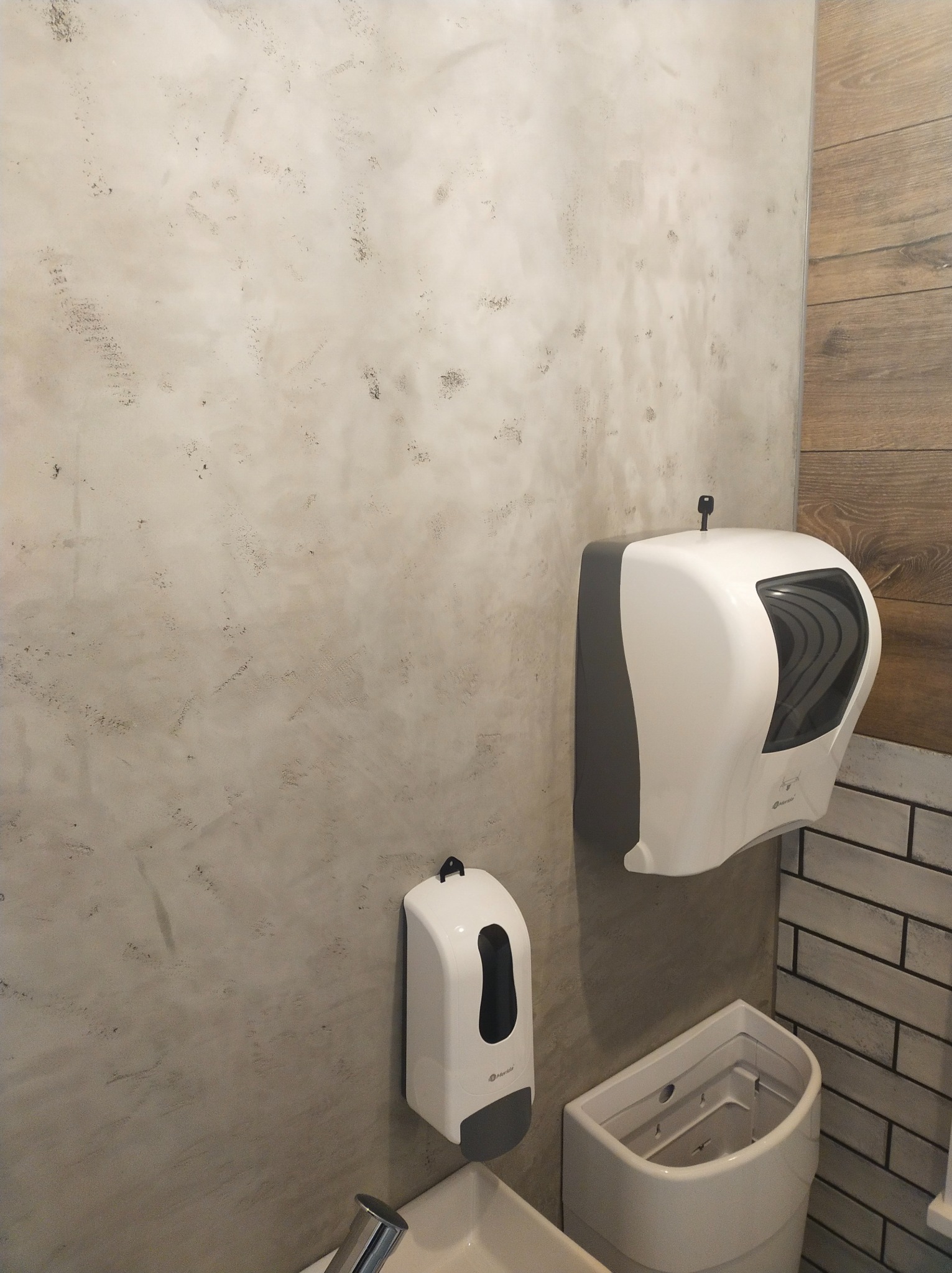 Ściana w łazience wykończona betonem architektonicznym, obok dozownik mydła w płynie i pojemnik na ręczniki papierowe.