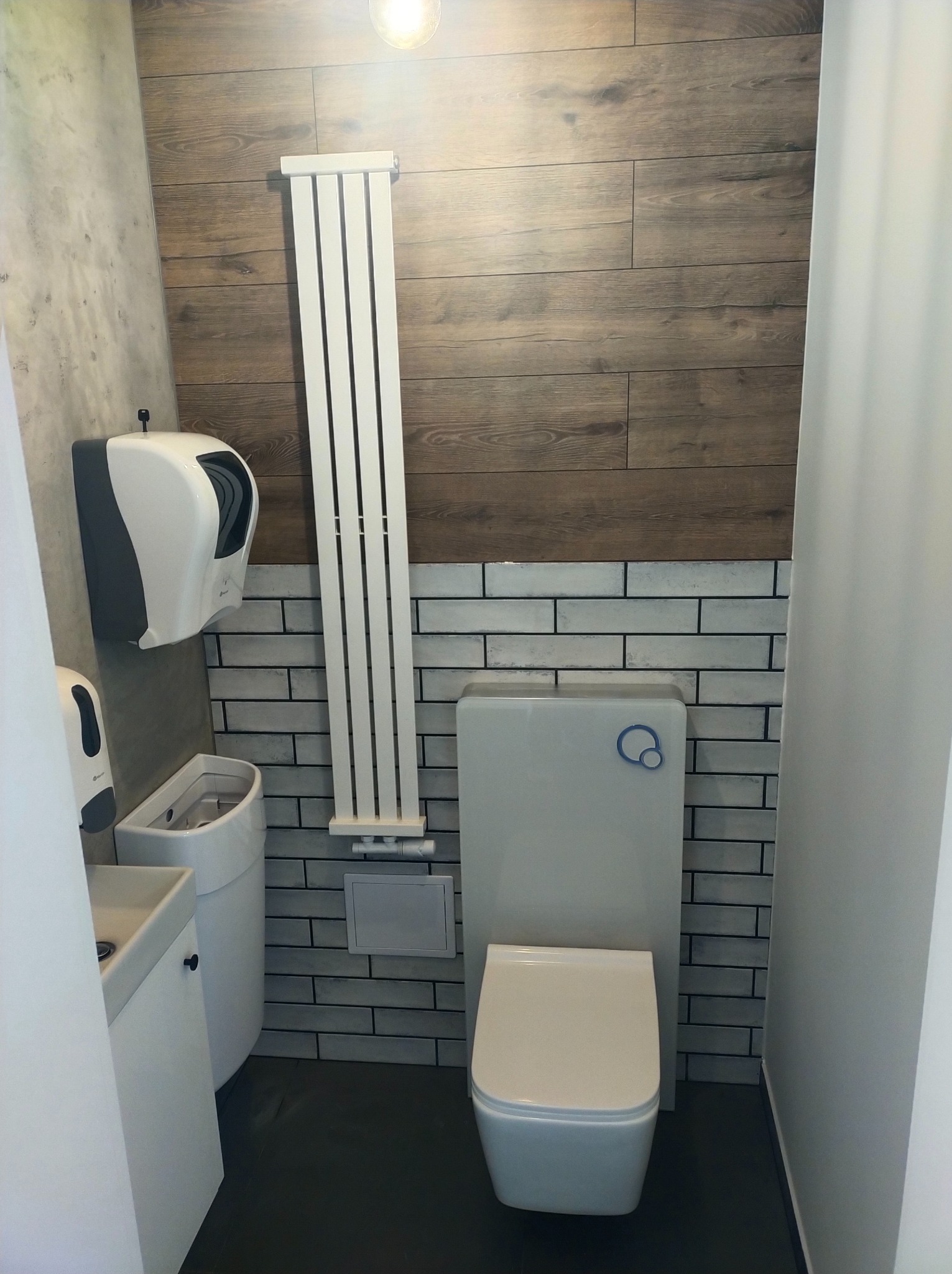 Nowoczesna łazienka w szaro-brązowej tonacji: drewnopodobne panele ścienne, białe płytki imitujące cegłę, pionowy grzejnik drabinkowy, wisząca toaleta z niebieskim akcentem oraz dozownik do mydła...