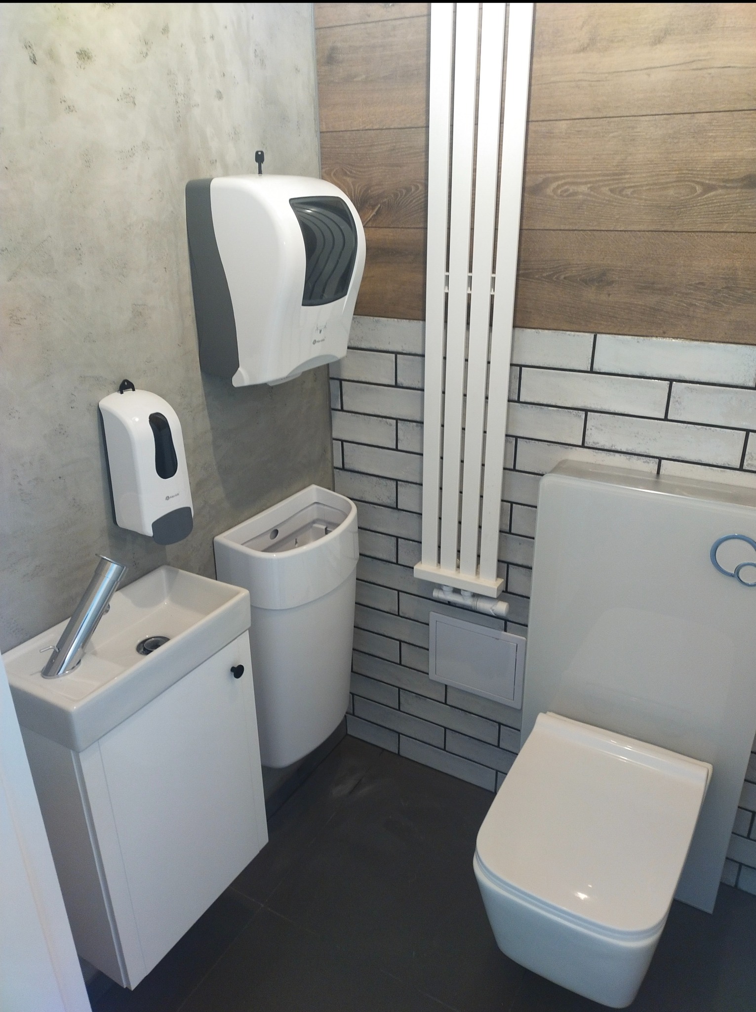 Nowoczesna, mała łazienka w szaro-białej tonacji z wiszącą toaletą, umywalką z szafką, grzejnikiem drabinkowym i dozownikami mydła, wykończona płytkami imitującymi cegłę.