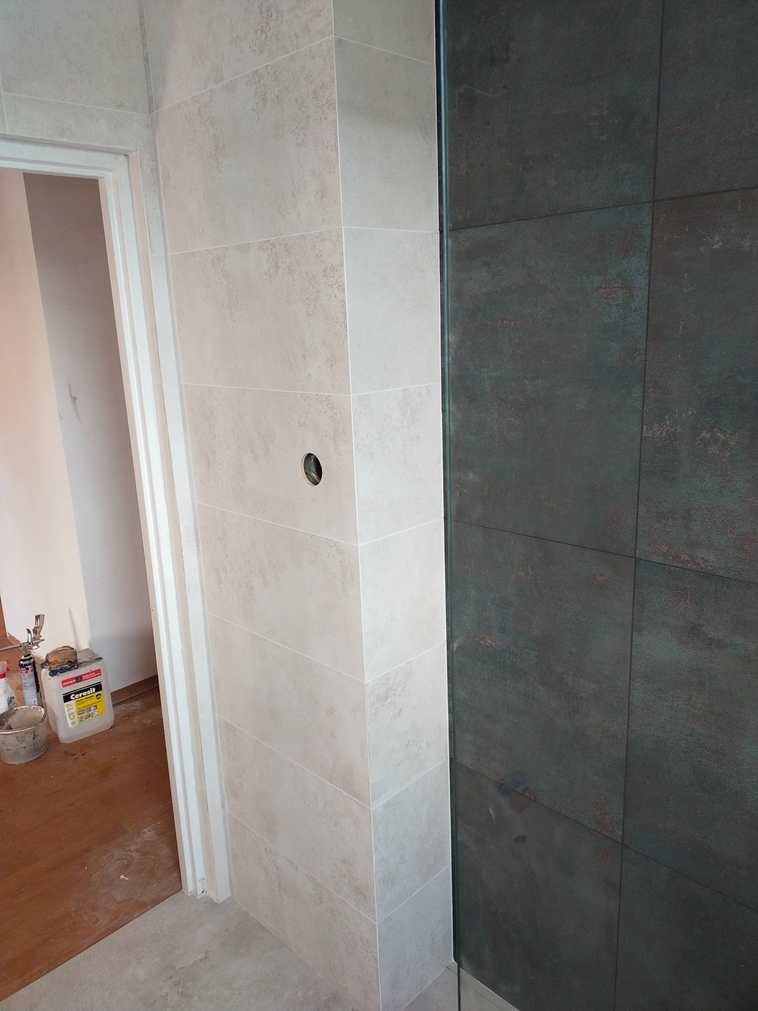 Wykończona ściana łazienki z jasnymi i ciemnymi płytkami, widoczny otwór na instalację, fragment drzwi i wiadro z klejem w tle.