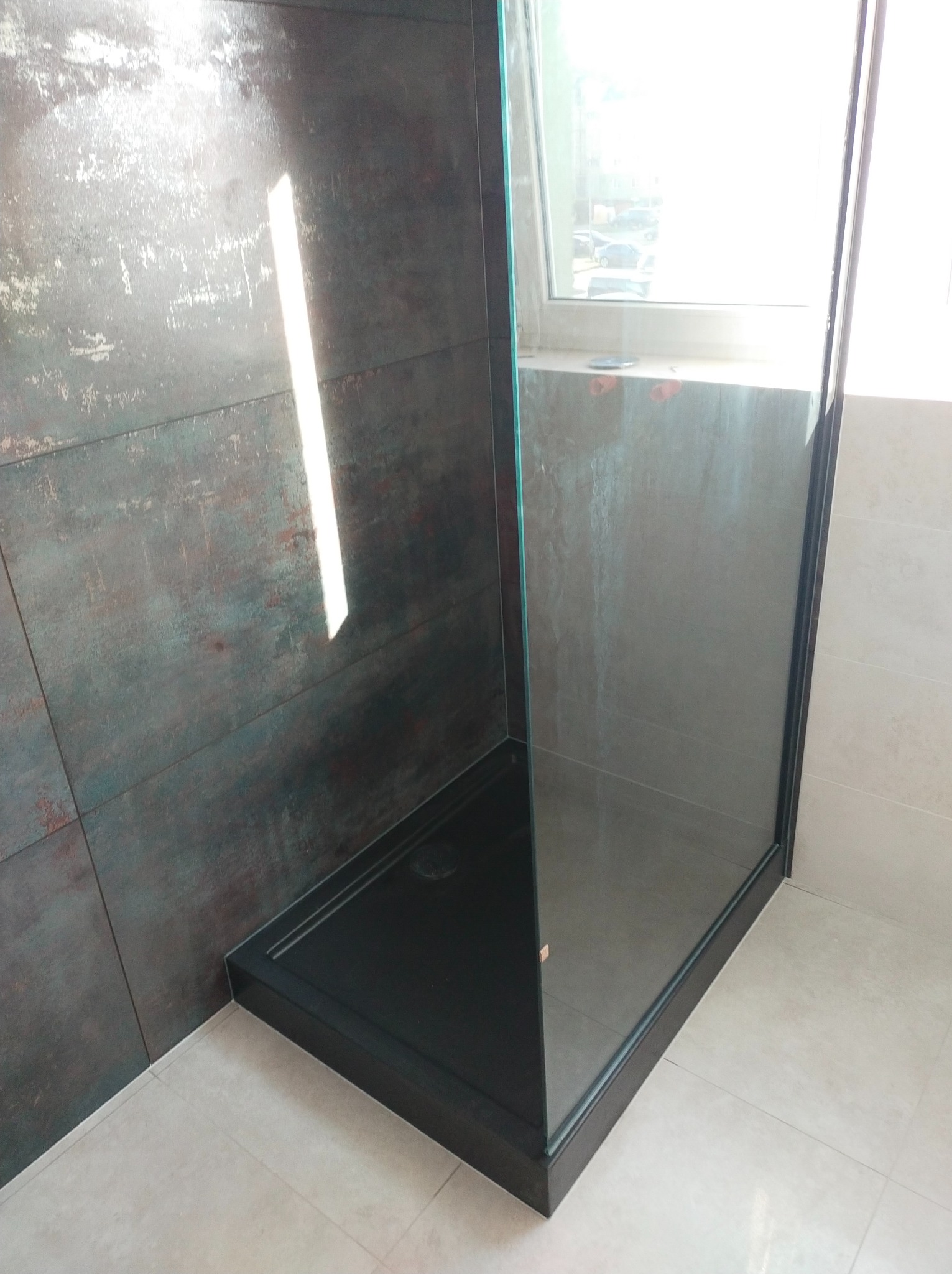 Kabina prysznicowa z czarnym brodzikiem i szklanymi ściankami, rustykalne ciemne płytki na ścianie, jasne płytki podłogowe, widok z niskiego kąta.