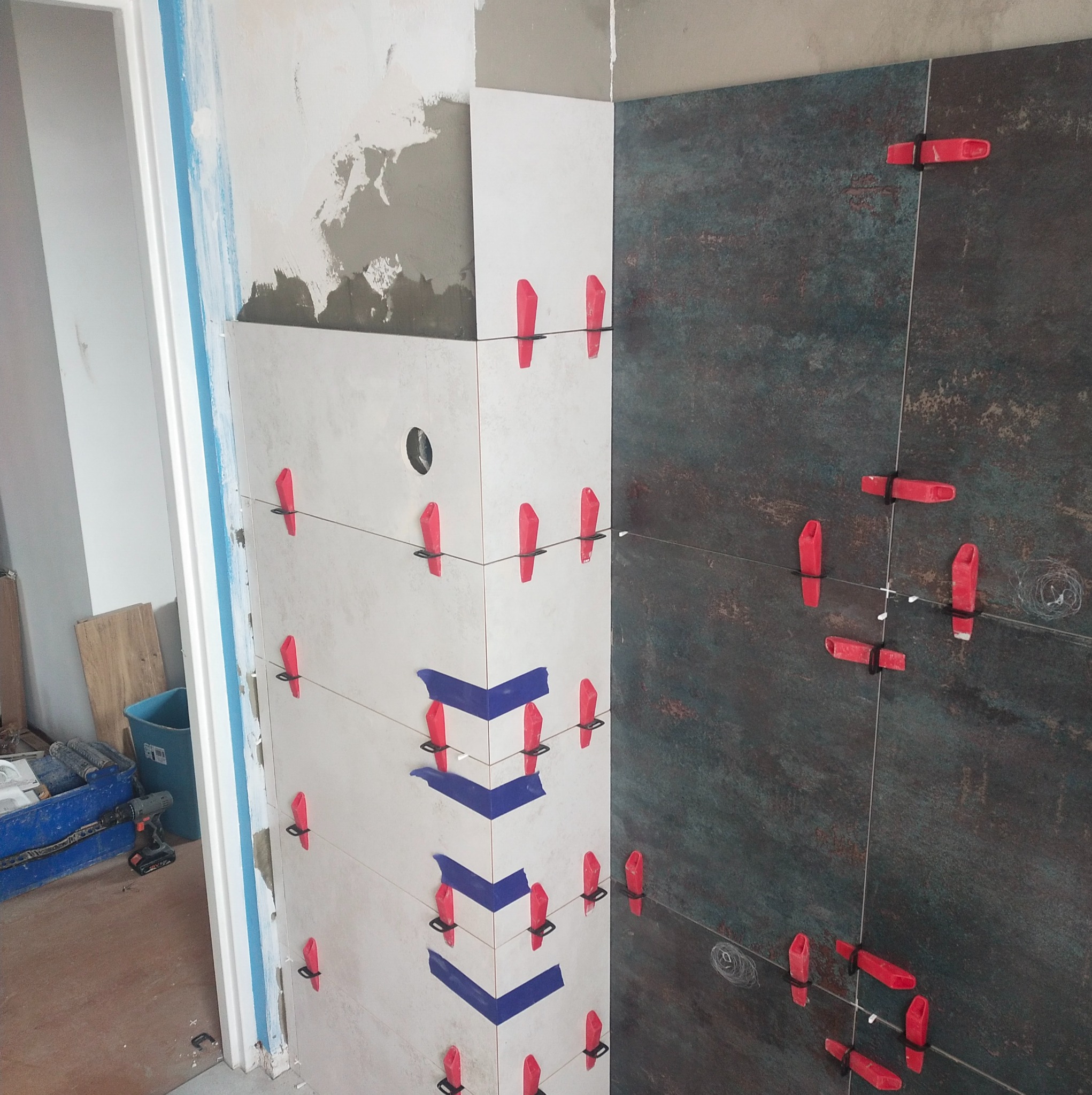 Układanie płytek w rogu pomieszczenia, użycie poziomowania z czerwonymi klipsami, niebieska taśma malarska wyznacza kąt, widoczne narzędzia w tle.