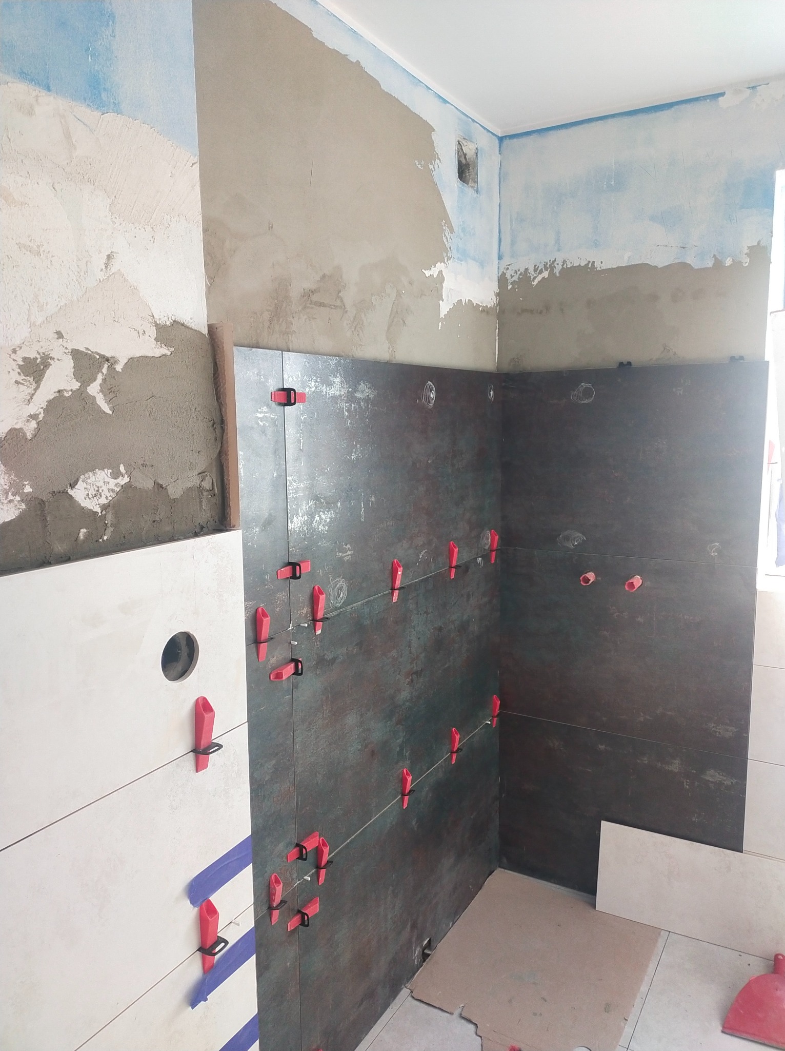 Montaż ciemnych płytek w narożniku łazienki, z użyciem czerwonych klipsów poziomujących, widoczne fragmenty surowej ściany i białe płytki z wyciętym otworem.