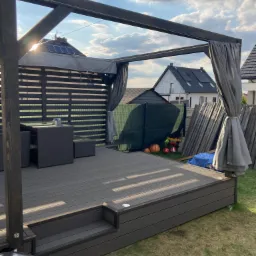 Zadaszona przestrzeń wypoczynkowa na zewnątrz z meblami ogrodowymi, drewnianą konstrukcją i zasłonami, widoczne elementy oświetlenia w podłodze, w tle domy z panelami słonecznymi na dachu.