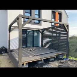 W trakcie budowy: Czarna drewniana konstrukcja z elementami tarasu kompozytowego, widoczne betonowe bloczki fundamentowe i narzędzia na deskach, na tle białego domu z szarymi oknami.