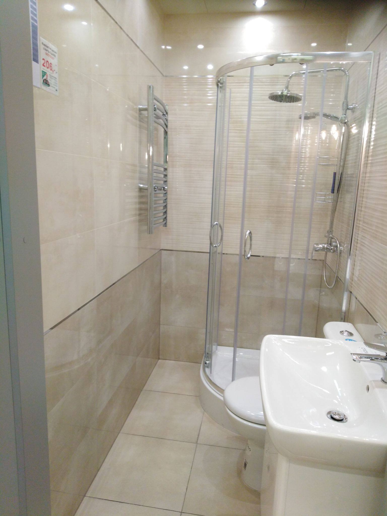 Wykończona łazienka w beżowych płytkach z kabiną prysznicową, grzejnikiem drabinkowym i białą umywalką na postumencie, widoczny plakat z ceną na ścianie.