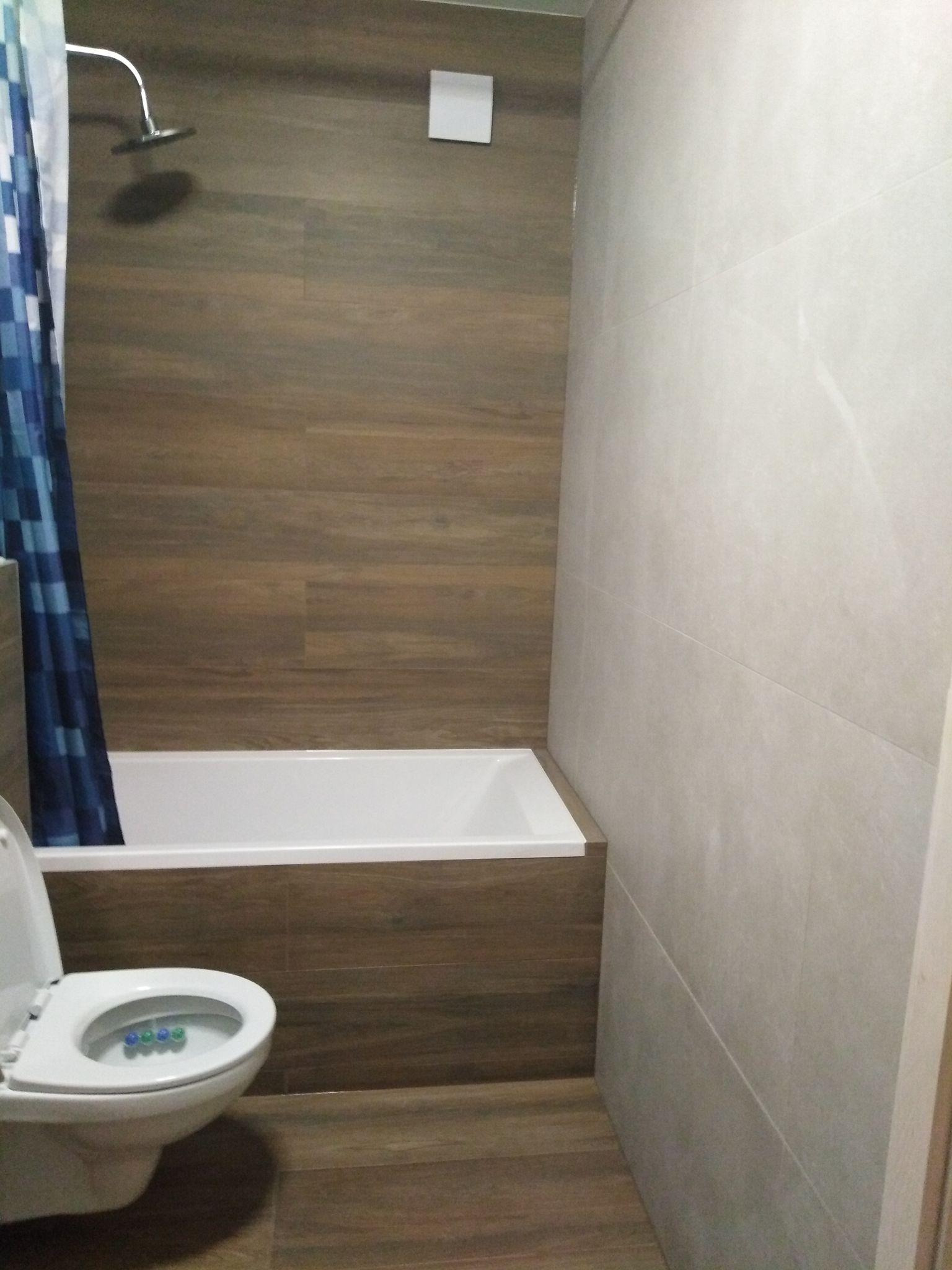 Wykończona łazienka z brązowymi płytkami imitującymi drewno na ścianie przy wannie, białą wanną, prysznicem i toaletą z odchyloną klapą, z widocznymi kostkami do WC.