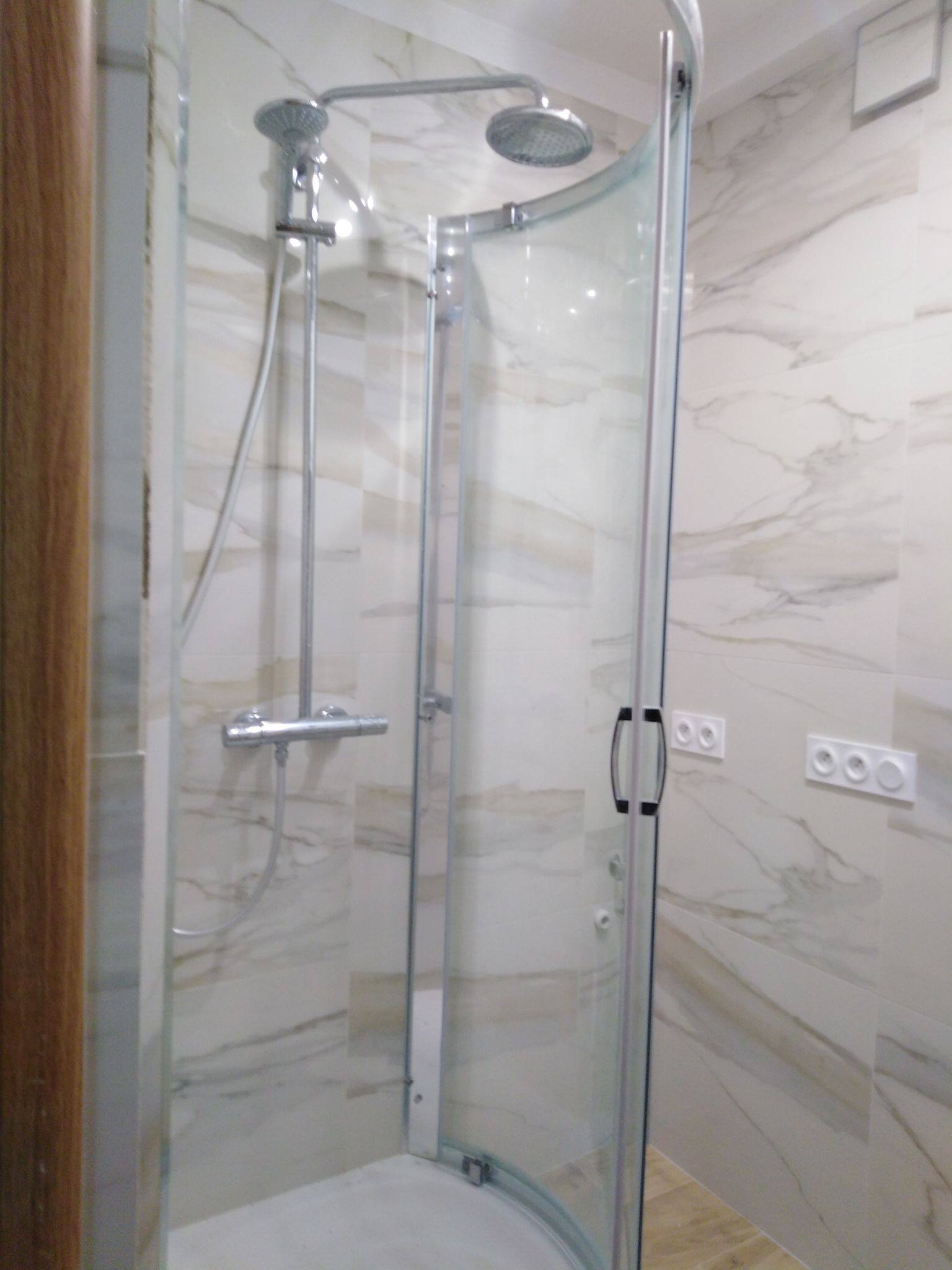 Nowoczesna kabina prysznicowa z deszczownicą, chromowana armatura, marmurowe płytki na ścianach, widoczne gniazdka elektryczne.