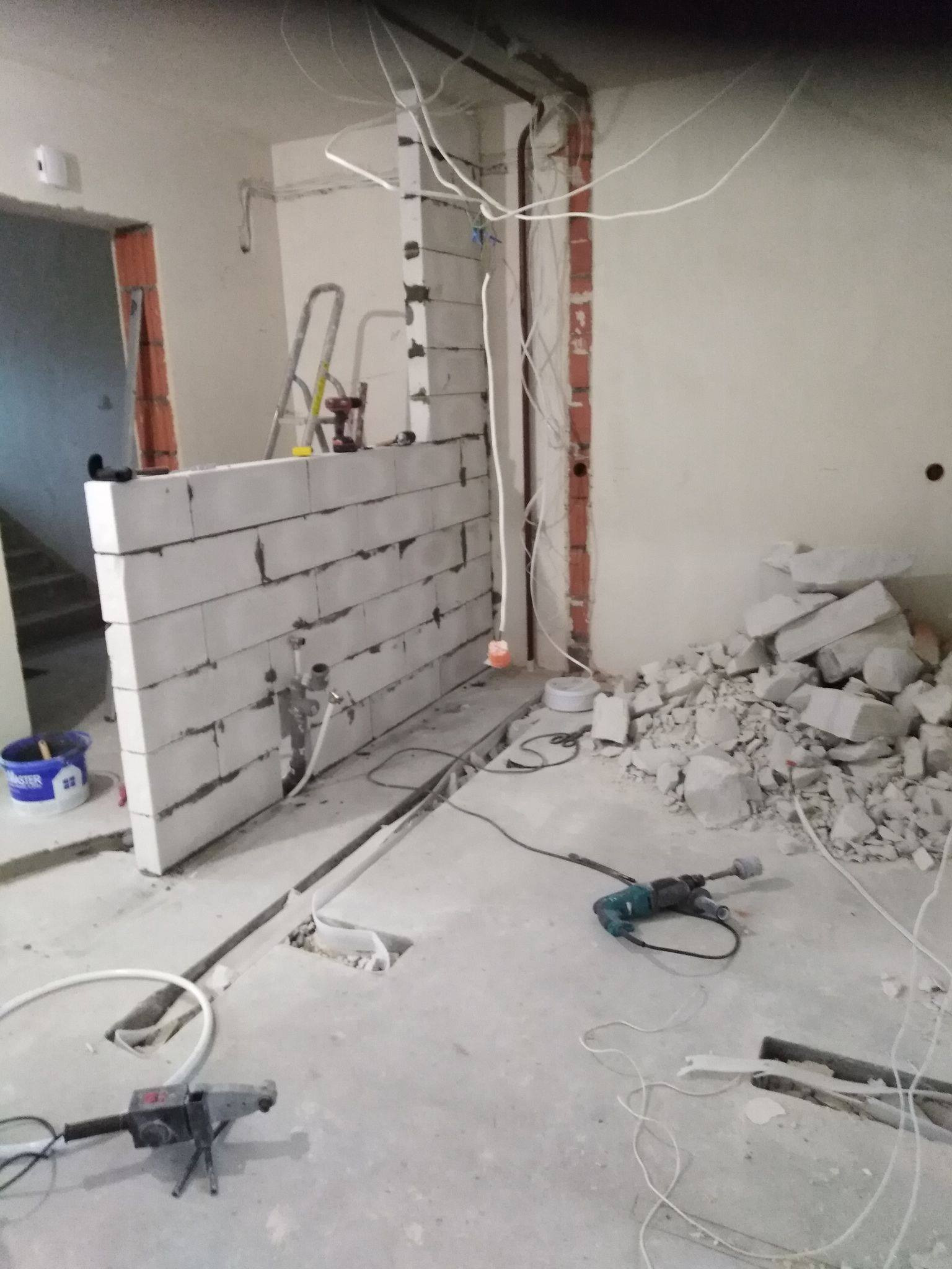 Wnętrze w trakcie prac remontowych: wzniesiona ściana z białych bloczków, odsłonięte przewody elektryczne, gruz, drabina i narzędzia budowlane na podłodze, widoczne otwory w podłodze na instalacje.
