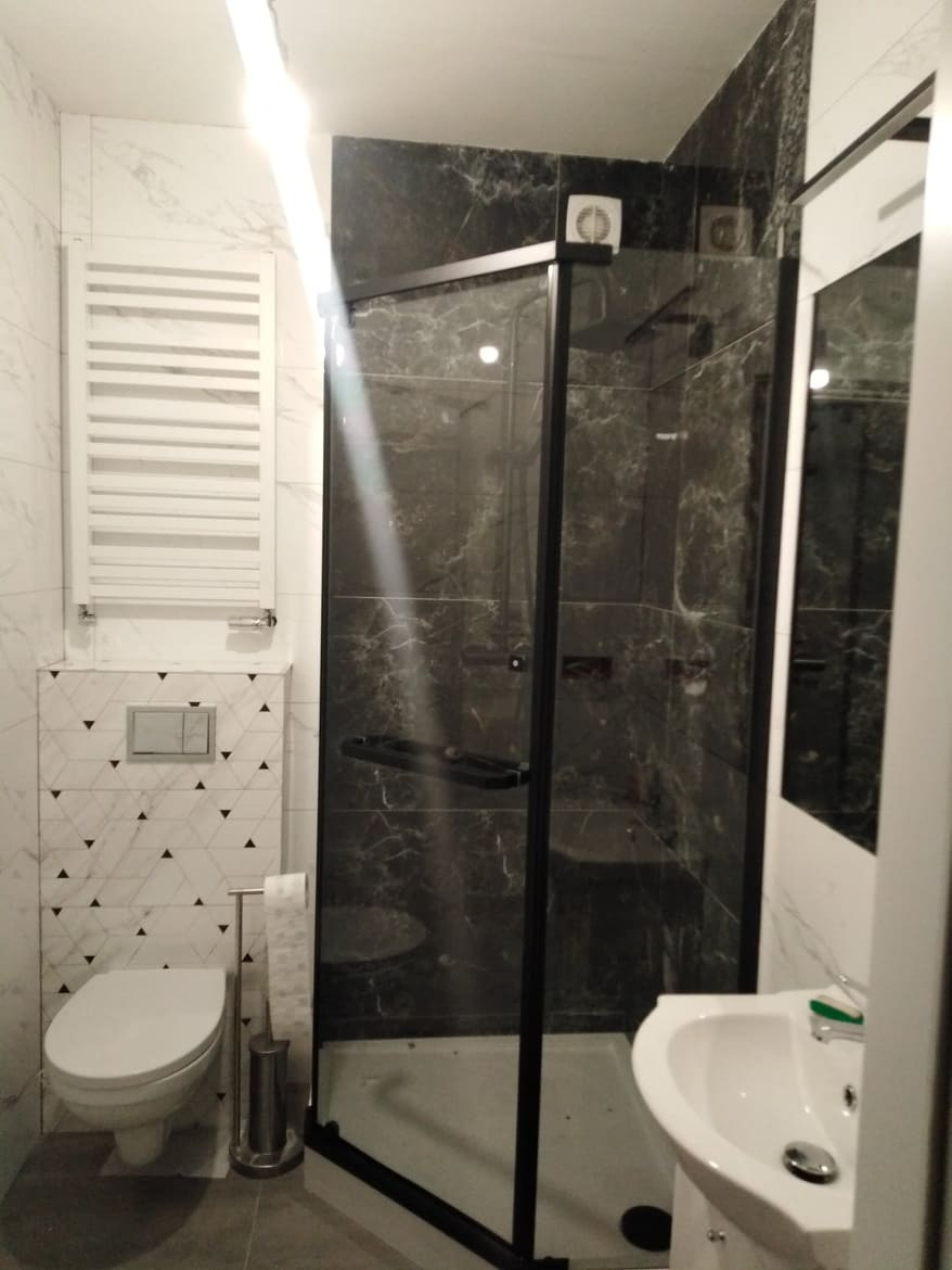 Nowoczesna łazienka z kabiną prysznicową typu walk-in z czarnymi ramami, marmurowe płytki na ścianach, podwieszana toaleta i grzejnik drabinkowy.