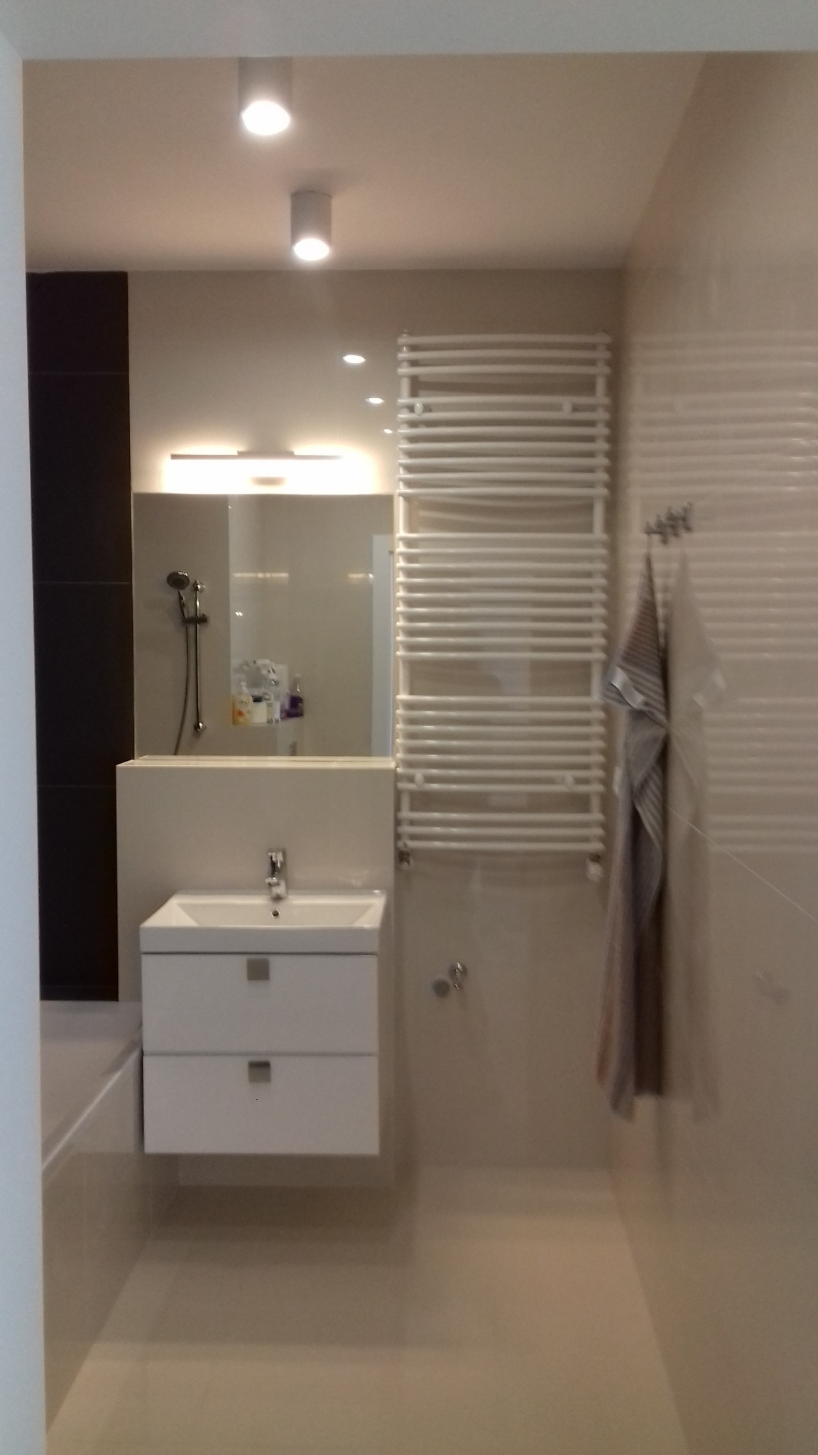 Nowoczesna łazienka z białymi płytkami, szafką pod umywalką z dwoma szufladami, grzejnikiem drabinkowym i ręcznikami w odcieniach szarości.