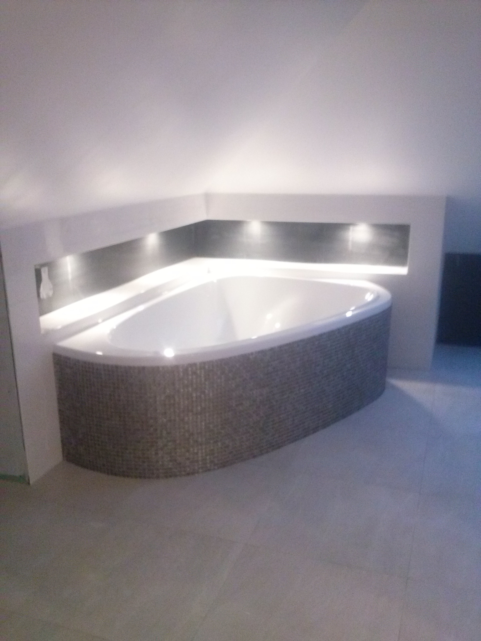 Nowoczesna wanna narożna obudowana mozaiką w odcieniach szarości, z oświetleniem punktowym w zabudowie ściennej, na jasnej podłodze.