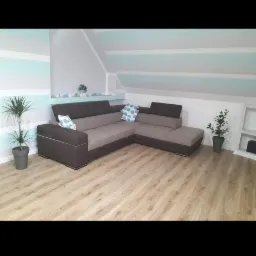 Nowoczesny narożnik w odcieniach brązu i szarości na drewnianej podłodze, w tle ściana w poziome, pastelowe pasy, dopełniony przez rośliny doniczkowe i dekoracyjne poduszki.