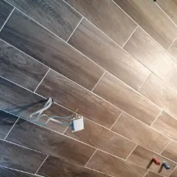 Ukośne ułożenie płytek imitujących drewno na ścianie łazienki, widoczne wystające kable i wtyczka w trakcie prac wykończeniowych.