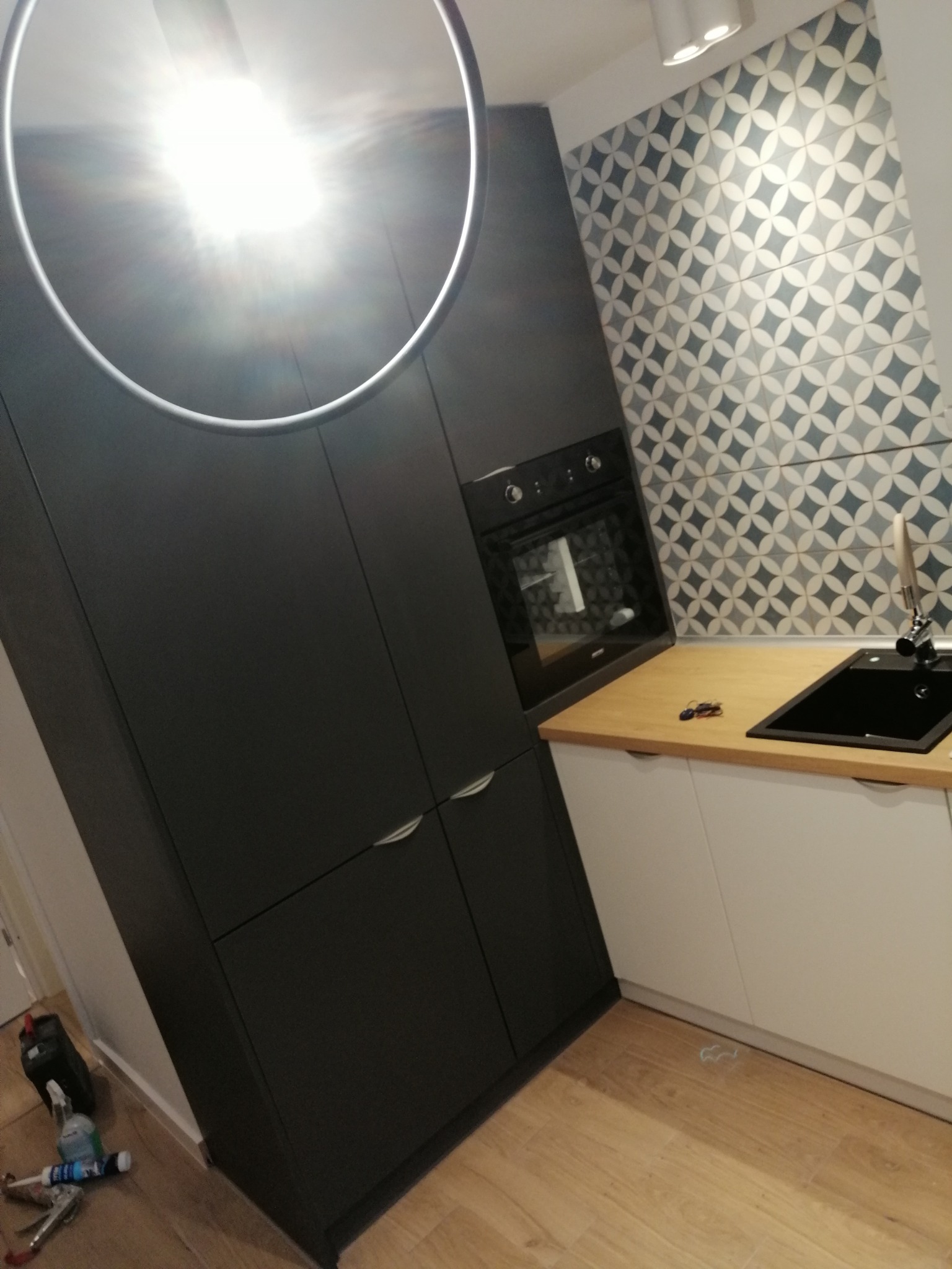 Nowoczesna kuchnia z ciemnoszarymi szafkami, drewnianym blatem, czarnym zlewozmywakiem i geometrycznymi płytkami na ścianie, oświetlona okrągłą lampą sufitową; na podłodze widoczne narzędzia...