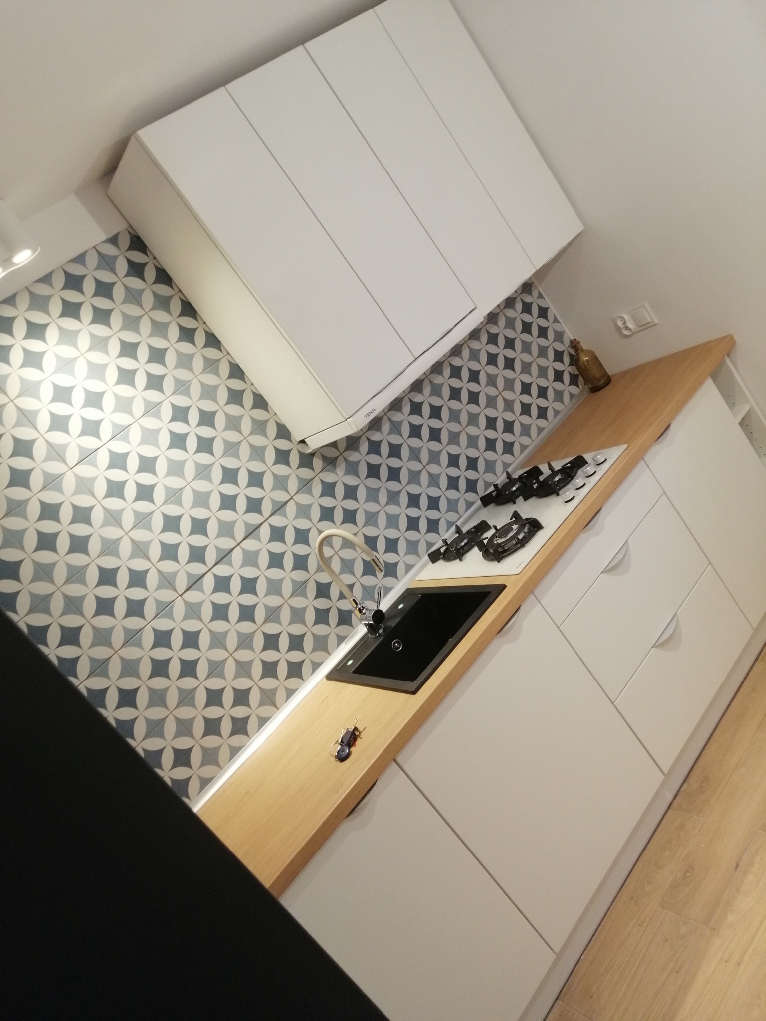 Wykończona kuchnia z białymi szafkami, drewnianym blatem, czarnym zlewem i płytą gazową na białym tle, ściana wyłożona dekoracyjnymi płytkami w geometryczny wzór.