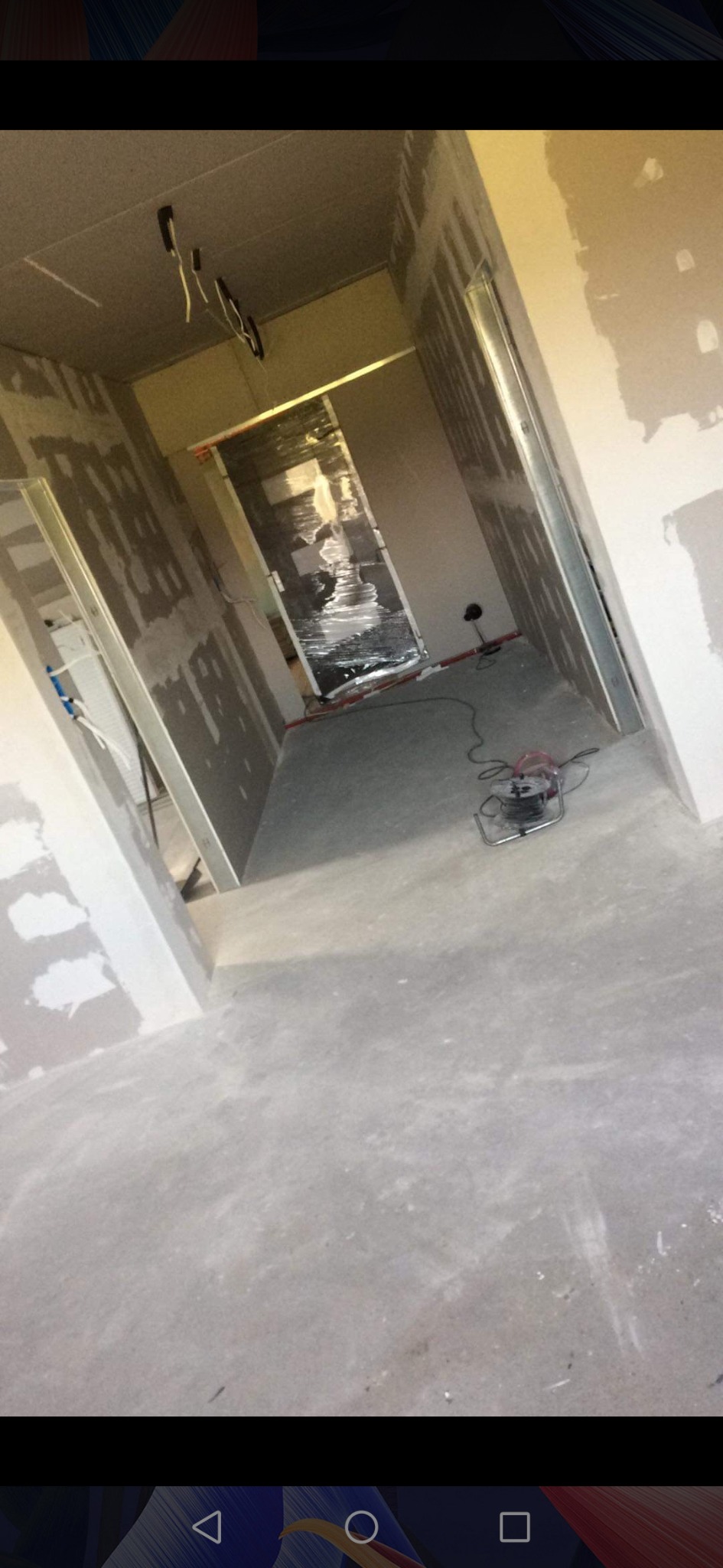 Wnętrze w trakcie prac wykończeniowych, widoczne płyty kartonowo-gipsowe na ścianach, surowy beton na podłodze i zabezpieczone folią drzwi, przewody elektryczne wystające z sufitu oraz zwinięty...