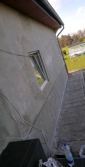 Ściana budynku po nałożeniu szarego tynku cementowo-wapiennego przed malowaniem, widoczne kable elektryczne, okno z uchylonym skrzydłem, rynna, w tle trawnik z altaną.