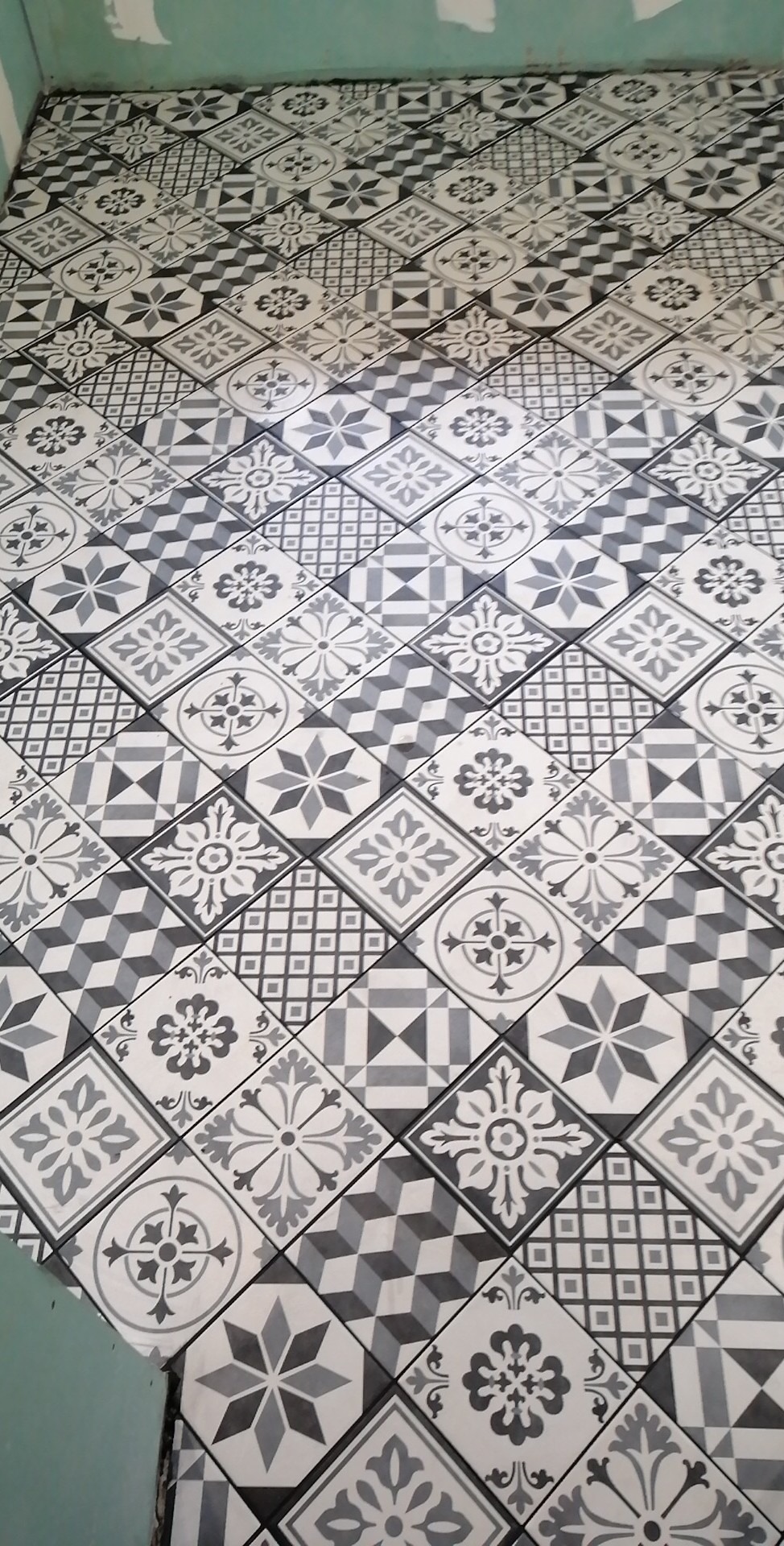 Ułożona pod kątem, biało-szara podłoga z płytek o zróżnicowanych wzorach geometrycznych i floralnych; widoczne niedociągnięcia przy ścianie.