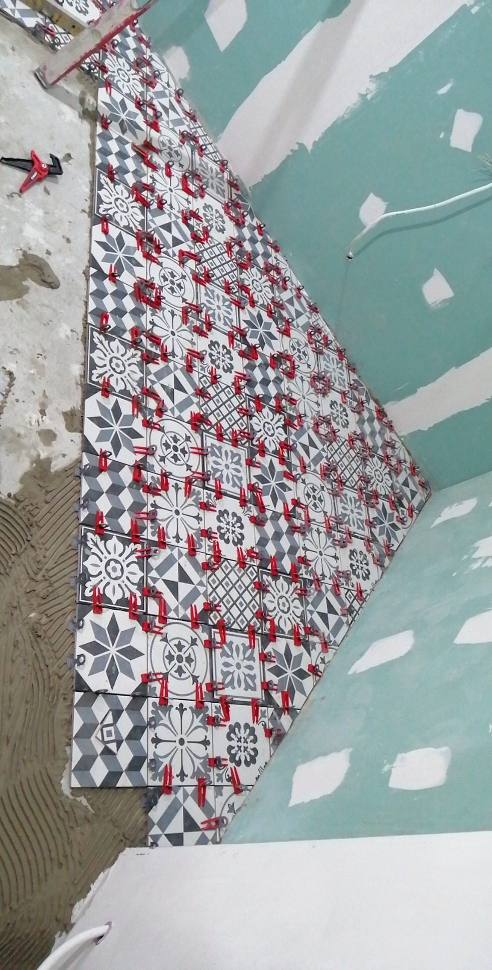 Ściana w trakcie wykańczania z szaro-białych płytek o wzorach geometrycznych i kwiatowych, z czerwonymi klipsami poziomującymi, widoczny fragment ściany z płyty kartonowo-gipsowej i resztki zaprawy.