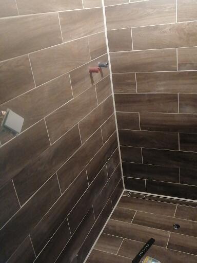 Świeżo wykafelkowany narożnik prysznica z brązowymi płytkami imitującymi drewno, widoczny odpływ liniowy i fragment butelki po środku czyszczącym.