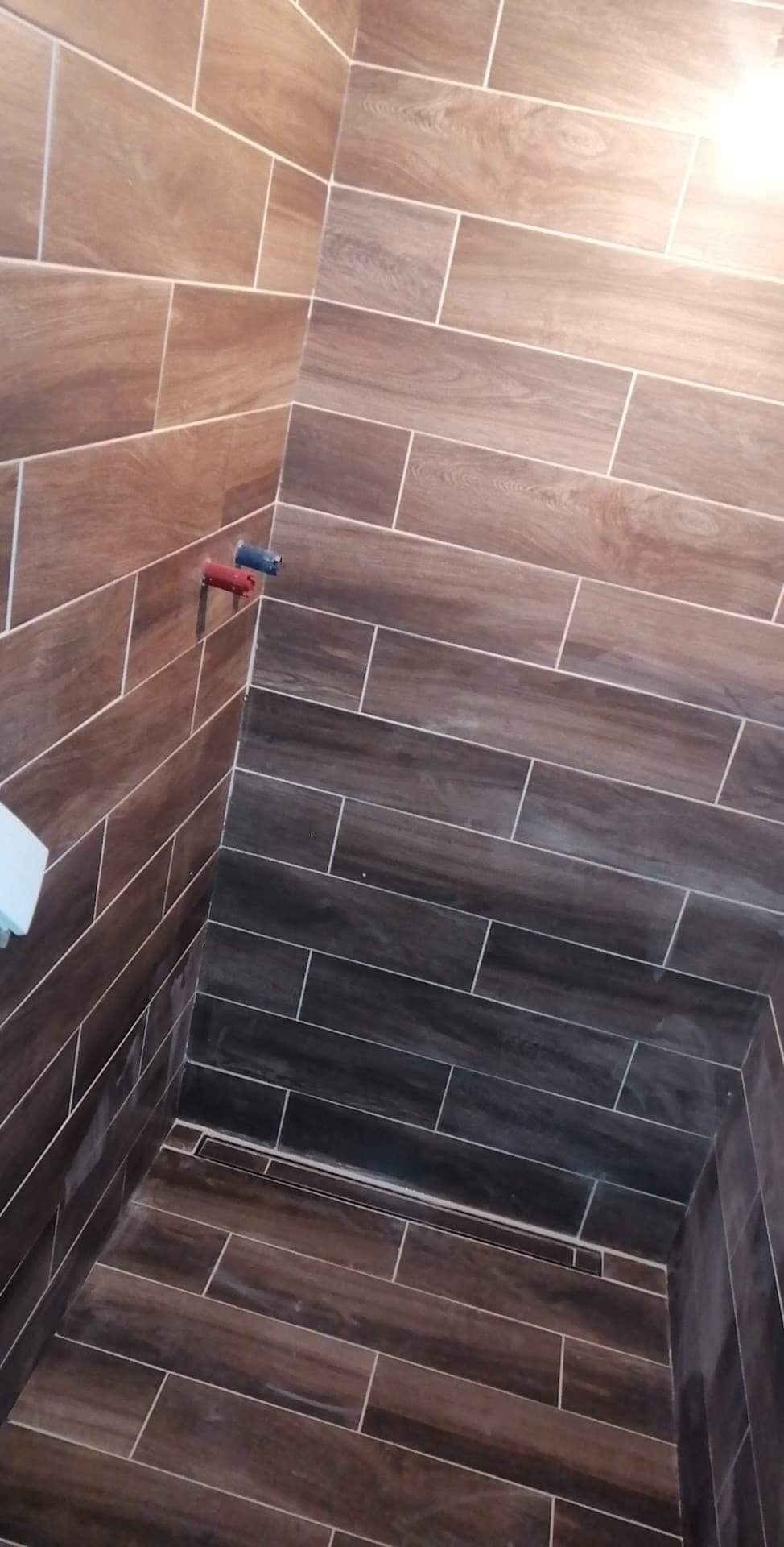 Wykończona kabina prysznicowa z brązowymi płytkami imitującymi drewno, widoczne rury z zaślepkami i odpływ liniowy w podłodze.