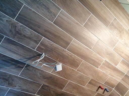 Ukośne ułożenie płytek imitujących drewno na ścianie łazienki, widoczne wystające kable i wtyczka w trakcie prac wykończeniowych.