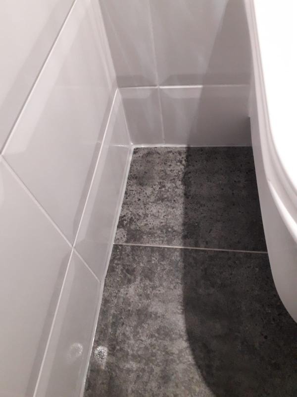 Fragment wykończonej łazienki z białymi, błyszczącymi płytkami ściennymi ułożonymi w geometryczny wzór i szarą, matową podłogą imitującą kamień, widoczny fragment toalety.