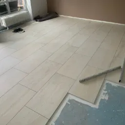 Układanie podłogi z płytek imitujących trawertyn, widoczny poziomica i fragment podłoża z pozostałościami kleju.