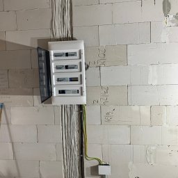 Olichem Jarosław Popławski - Rozdzielnica elektryczna z otwartymi drzwiczkami zamontowana na ścianie z białych bloczków betonowych, widoczne kable prowadzone w bruzdach, surowy stan wykończenia.