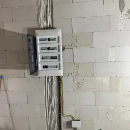 Rozdzielnica elektryczna z otwartymi drzwiczkami zamontowana na ścianie z białych bloczków betonowych, widoczne kable prowadzone w bruzdach, surowy stan wykończenia.