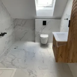 Nowoczesna łazienka na poddaszu z marmurowymi płytkami na ścianach i podłodze, drewniana szafka pod umywalką częściowo zabezpieczona folią bąbelkową, wisząca toaleta i okno dachowe wpuszczające...