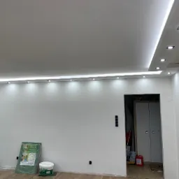 Wnętrze w trakcie prac wykończeniowych, widoczny sufit podwieszany z oświetleniem LED, ściany częściowo przygotowane do malowania, puszki elektryczne, wiadro z farbą i folia ochronna na podłodze.
