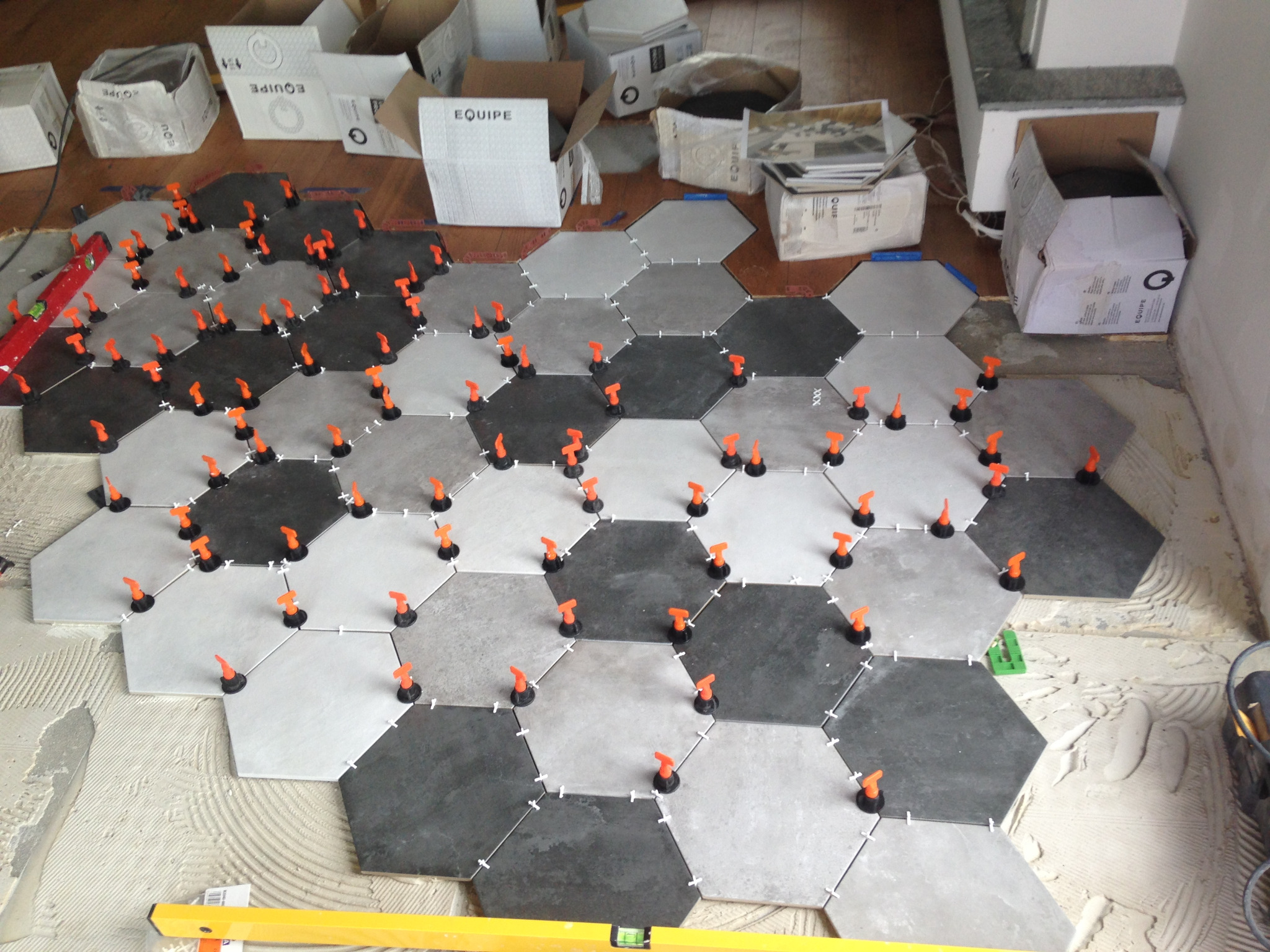 Układanie heksagonalnych płytek podłogowych w odcieniach szarości z użyciem pomarańczowych klinów poziomujących, widoczne pudełka po płytkach i poziomica.