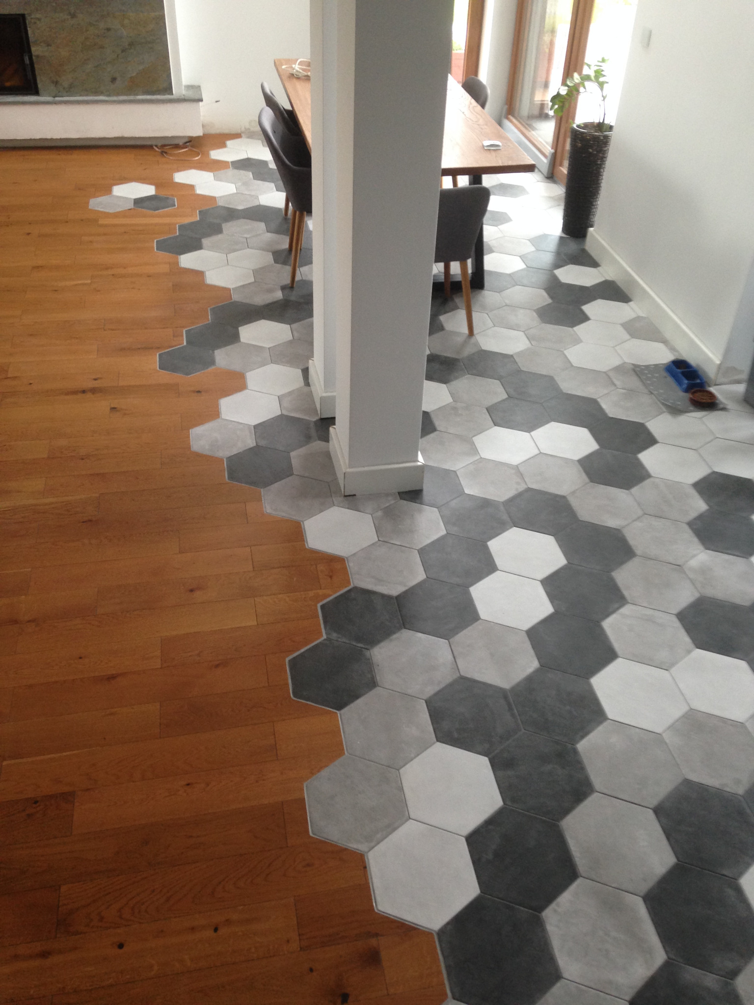 Podłoga łącząca drewniane panele z szarymi i białymi heksagonalnymi płytkami, widok na jadalnię z białymi ścianami i doniczką z rośliną.