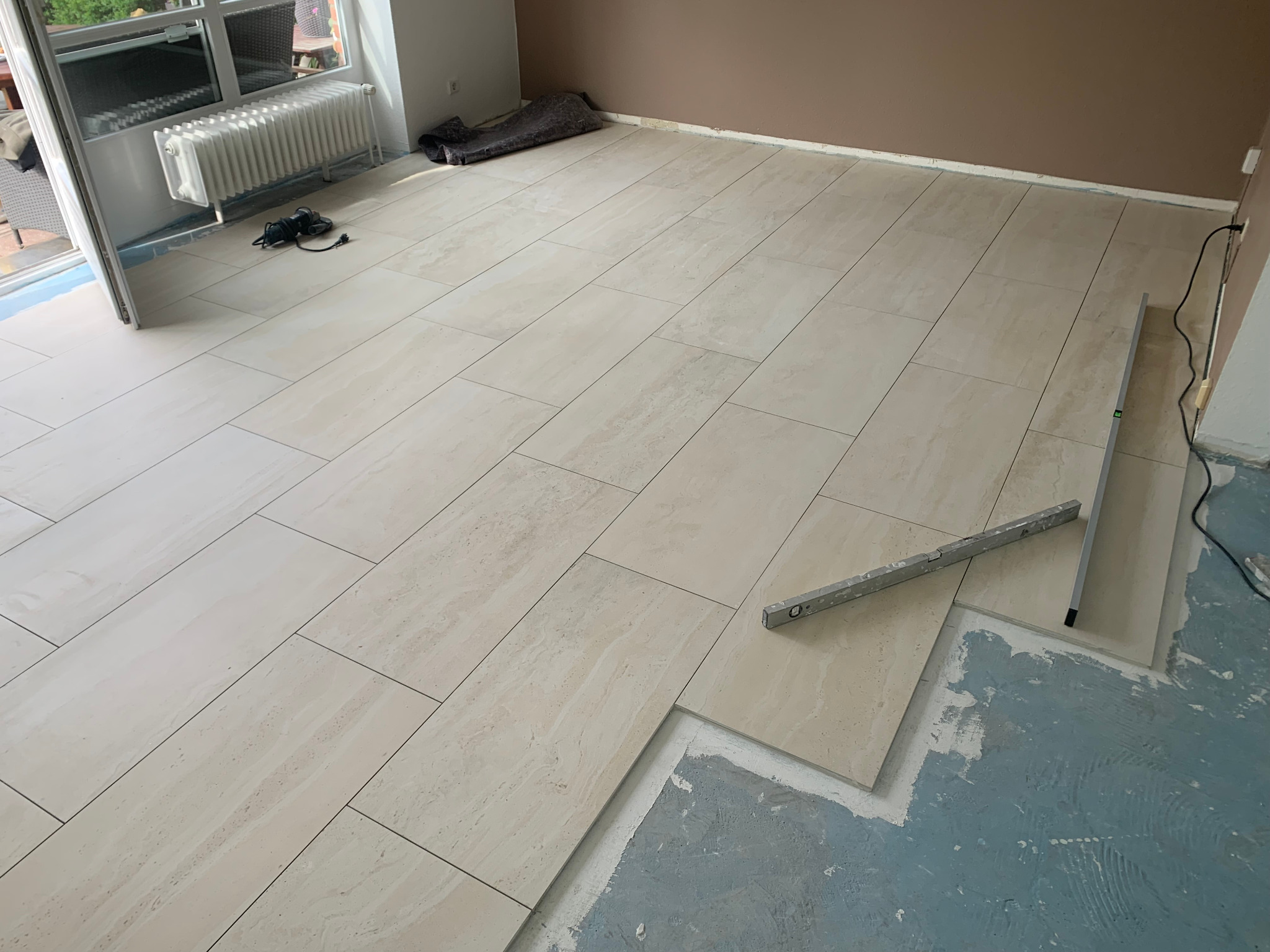 Układanie podłogi z płytek imitujących trawertyn, widoczny poziomica i fragment podłoża z pozostałościami kleju.