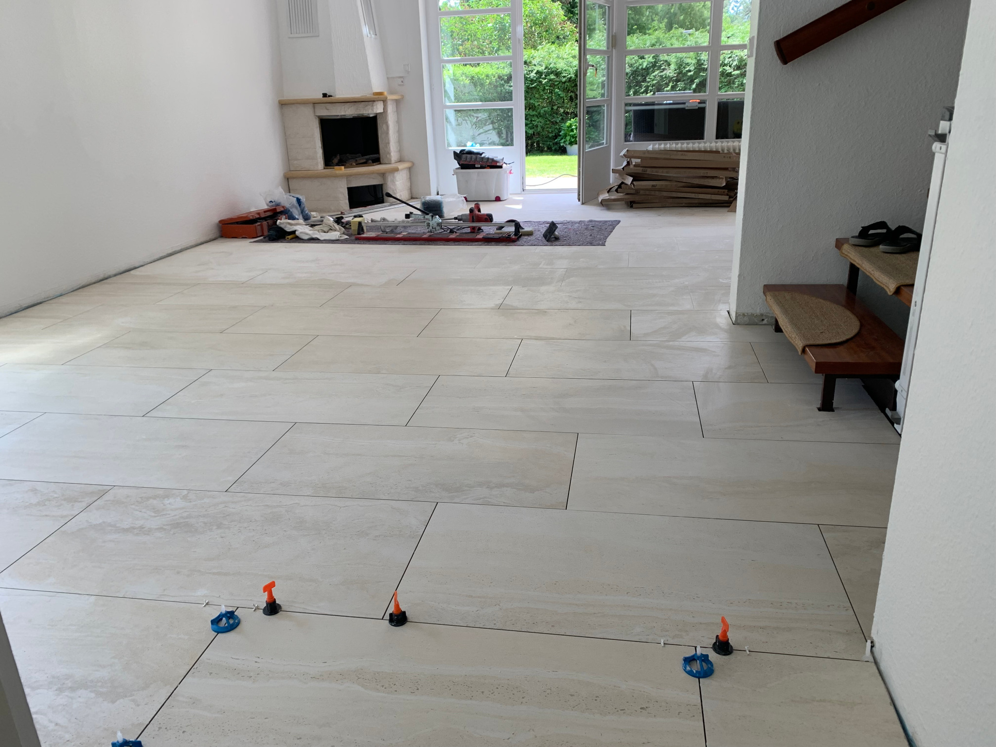 Jasne, prostokątne płytki podłogowe układane z użyciem systemów poziomowania, widoczne narzędzia i materiały budowlane w tle, otwarte drzwi tarasowe w jasnym wnętrzu.