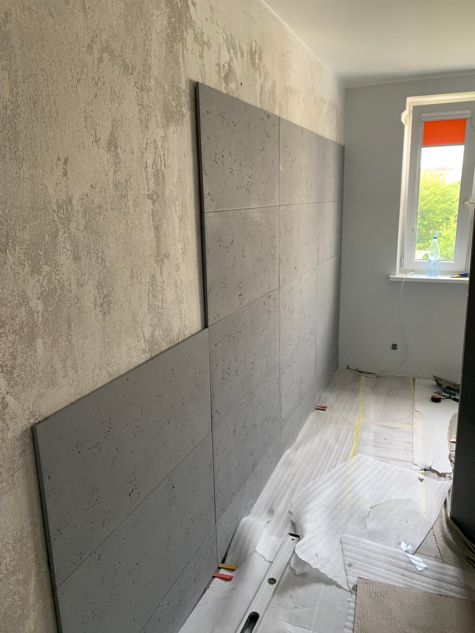 Montaż szarych paneli dekoracyjnych imitujących beton na ścianie w łazience, widoczne poziomica, butelka wody na parapecie i częściowo zakryta podłoga.