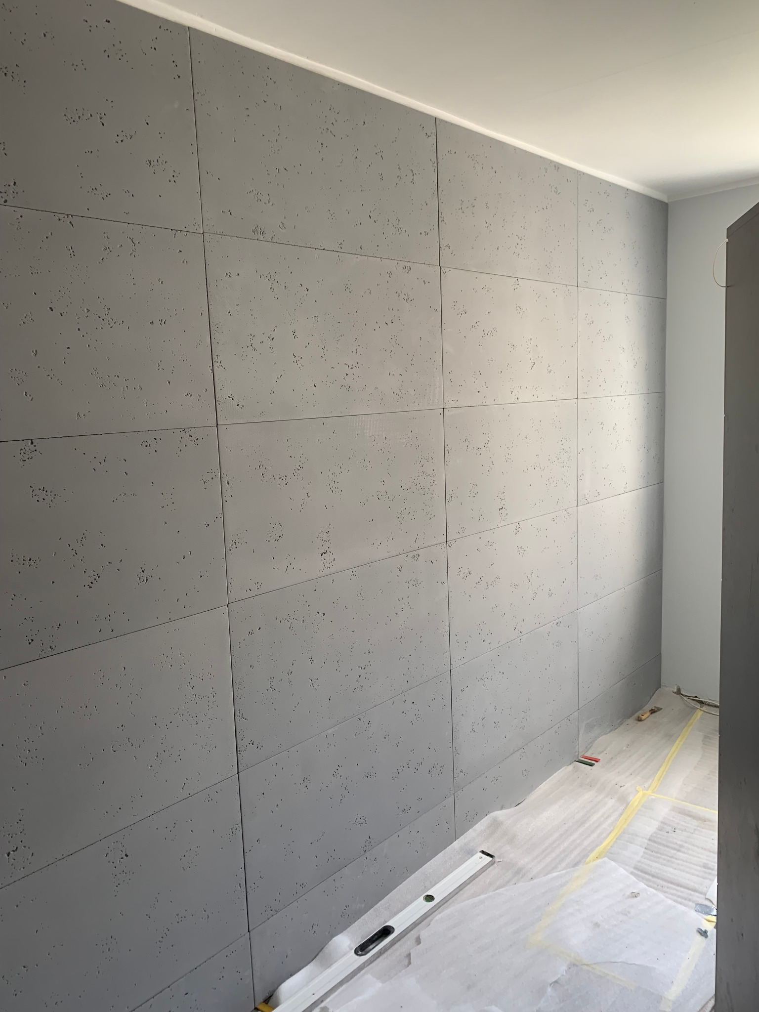 Ściana wyłożona szarymi panelami imitującymi beton architektoniczny, widoczny poziomica i folia ochronna na podłodze, przygotowanie do dalszych prac.