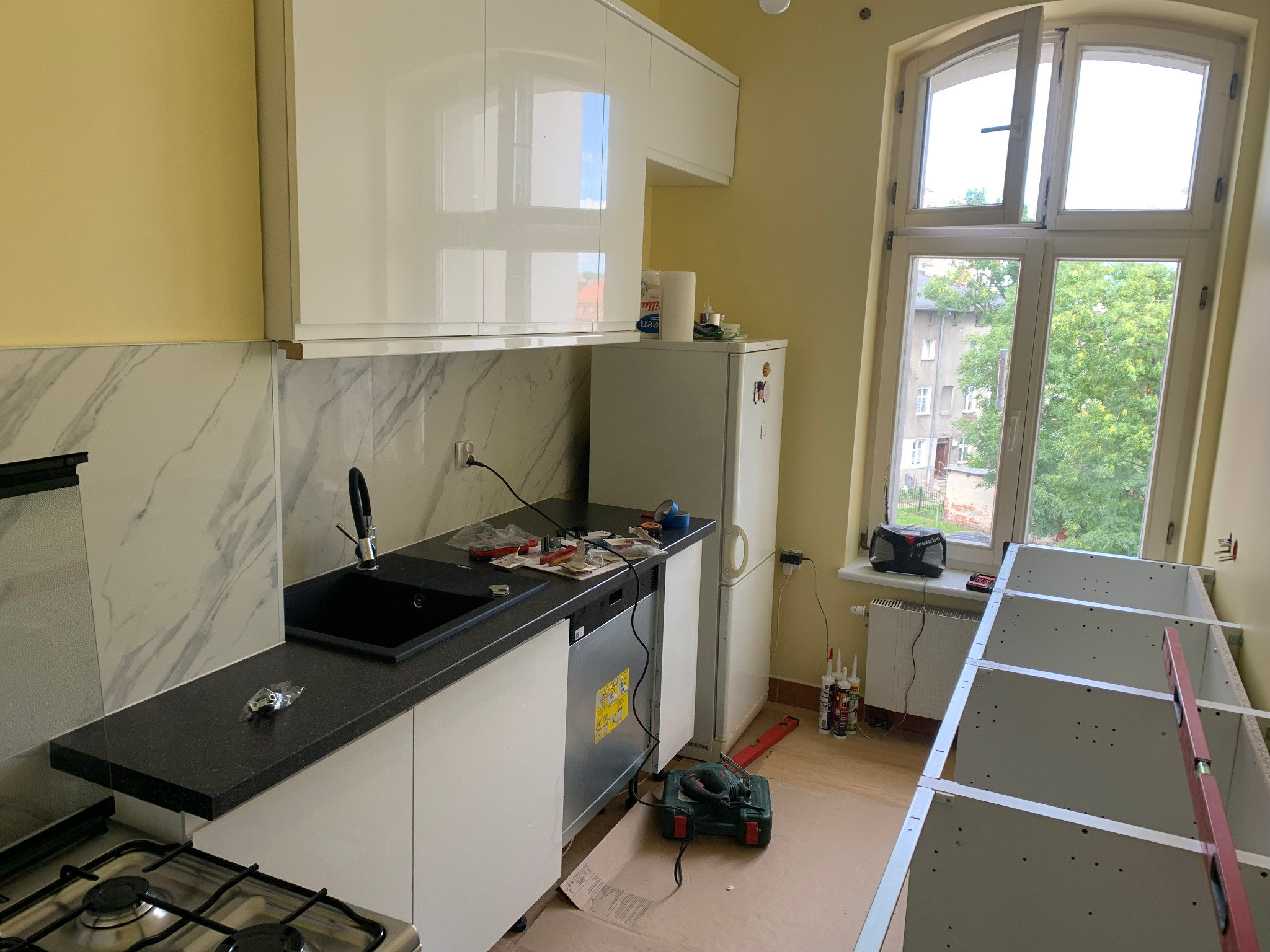 Wnętrze kuchni w trakcie montażu: białe, lakierowane szafki wiszące i dolne z czarnym blatem, czarny zlew, biała lodówka, okno z widokiem na zieleń, narzędzia rozłożone na blacie, częściowo...