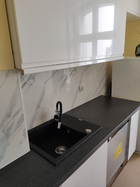 Czarne zlewozmywak z baterią na blacie w kuchni z białymi szafkami i marmurowym panelem ściennym.