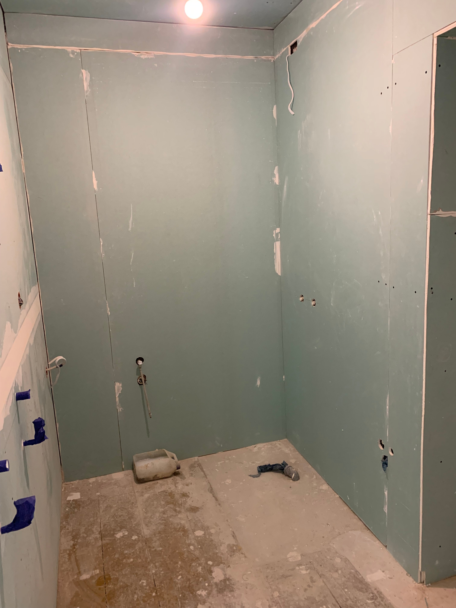 Łazienka w trakcie remontu: ściany wyłożone płytami gipsowo-kartonowymi, widoczne instalacje wodne zabezpieczone niebieską folią, podłoga przygotowana do dalszych prac, narzędzia i wiadro na podłodze.