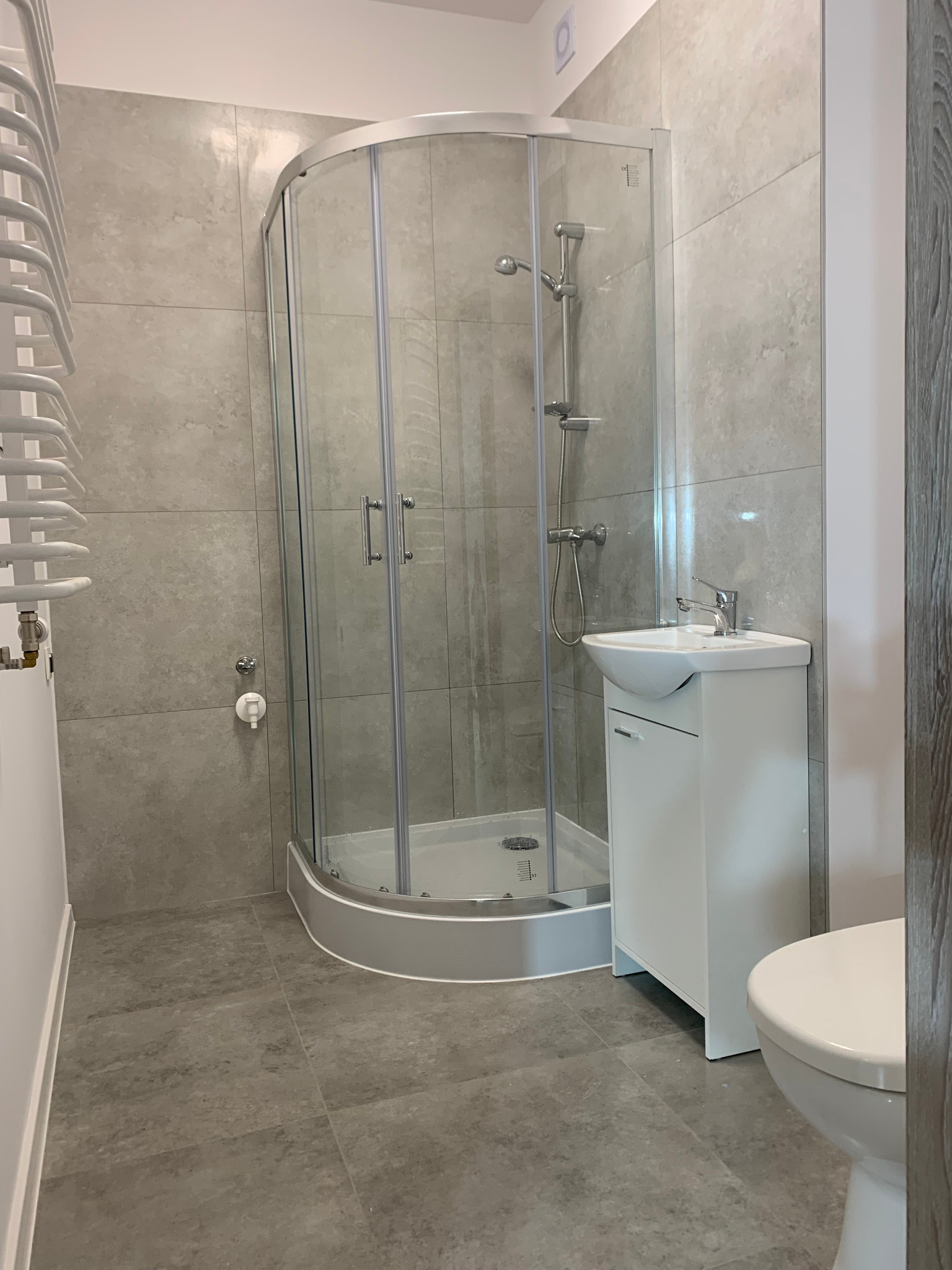 Nowoczesna łazienka z szarą glazurą, kabiną prysznicową narożną z przezroczystego szkła, białą umywalką z szafką oraz grzejnikiem drabinkowym po lewej stronie.