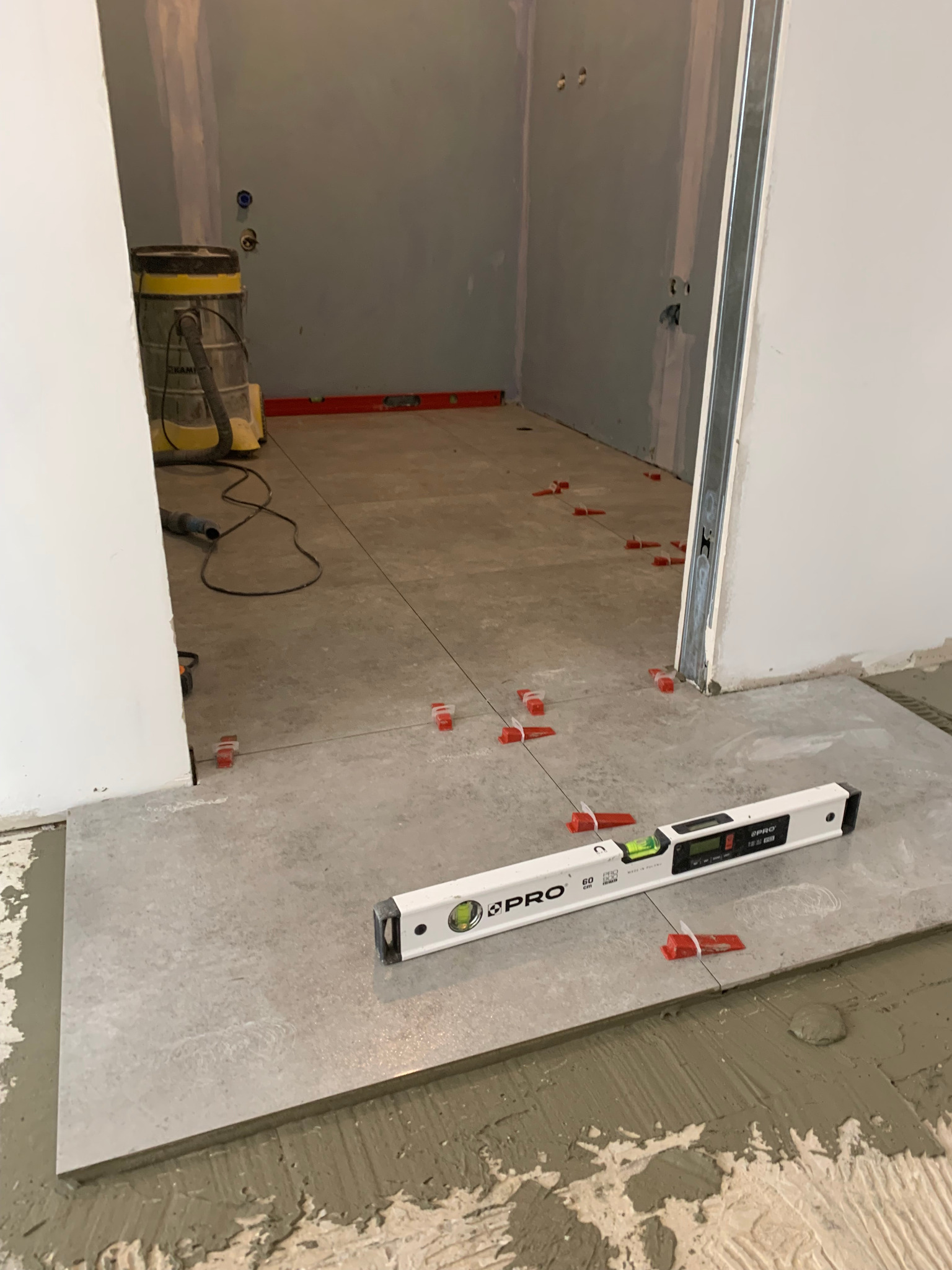 Szara płytka podłogowa układana z użyciem poziomicy i czerwonych klinów dystansowych, widoczny odkurzacz przemysłowy w tle, surowe ściany przygotowane do wykończenia.