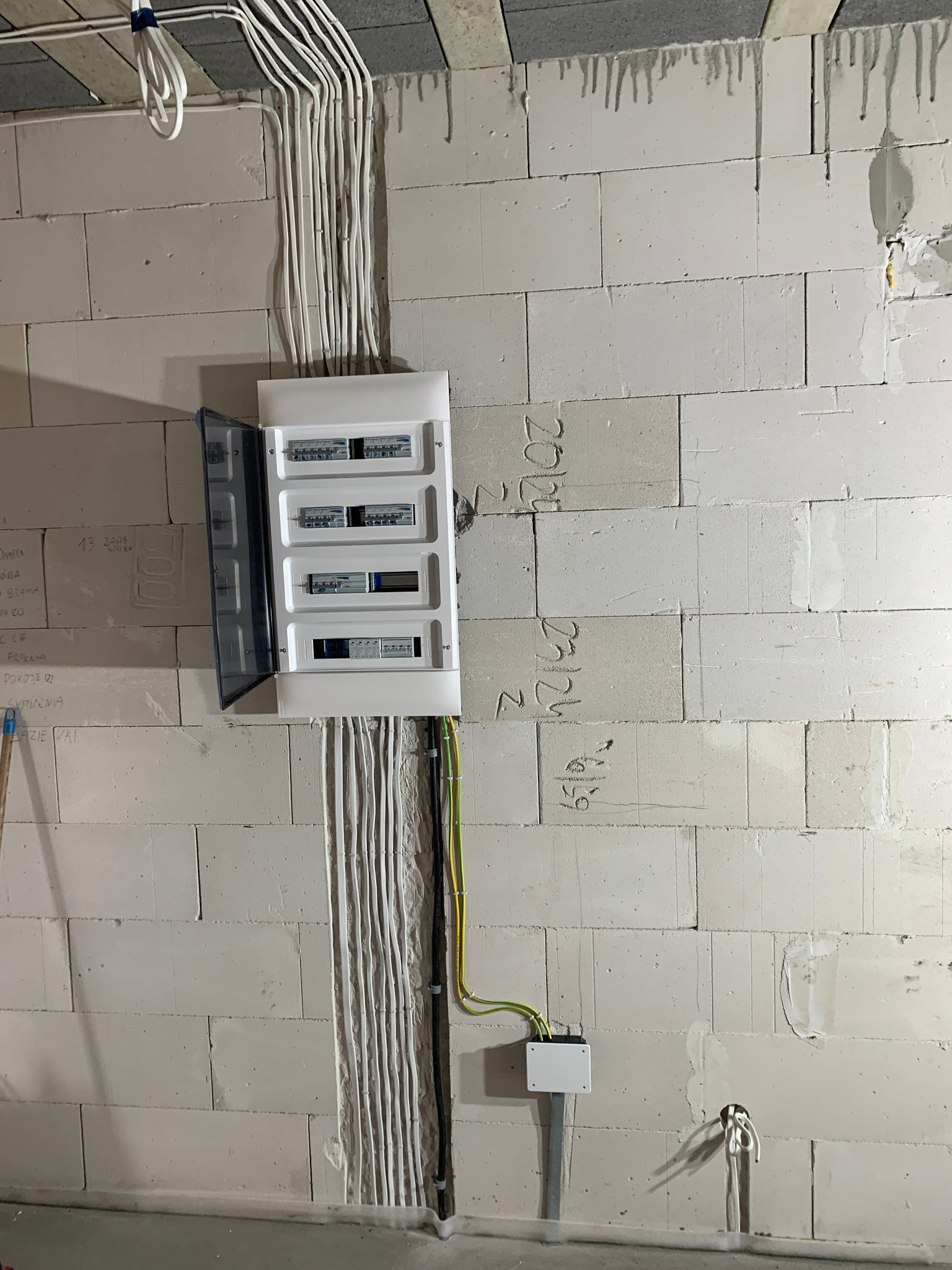Rozdzielnica elektryczna z otwartymi drzwiczkami zamontowana na ścianie z białych bloczków betonowych, widoczne kable prowadzone w bruzdach, surowy stan wykończenia.