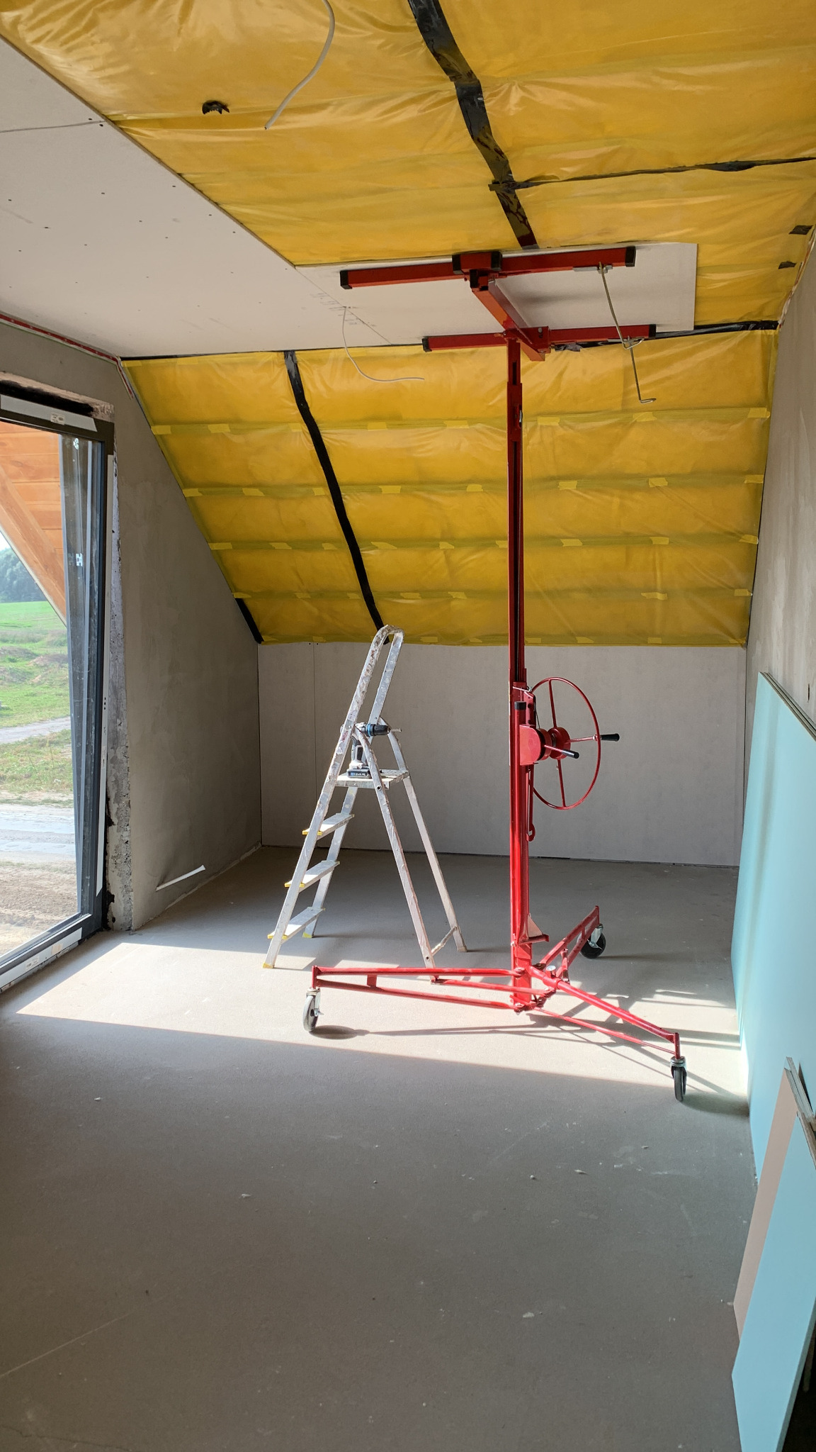 Montaż płyty gipsowo-kartonowej na poddaszu z użyciem podnośnika, widoczna drabina i folia izolacyjna na skosach.