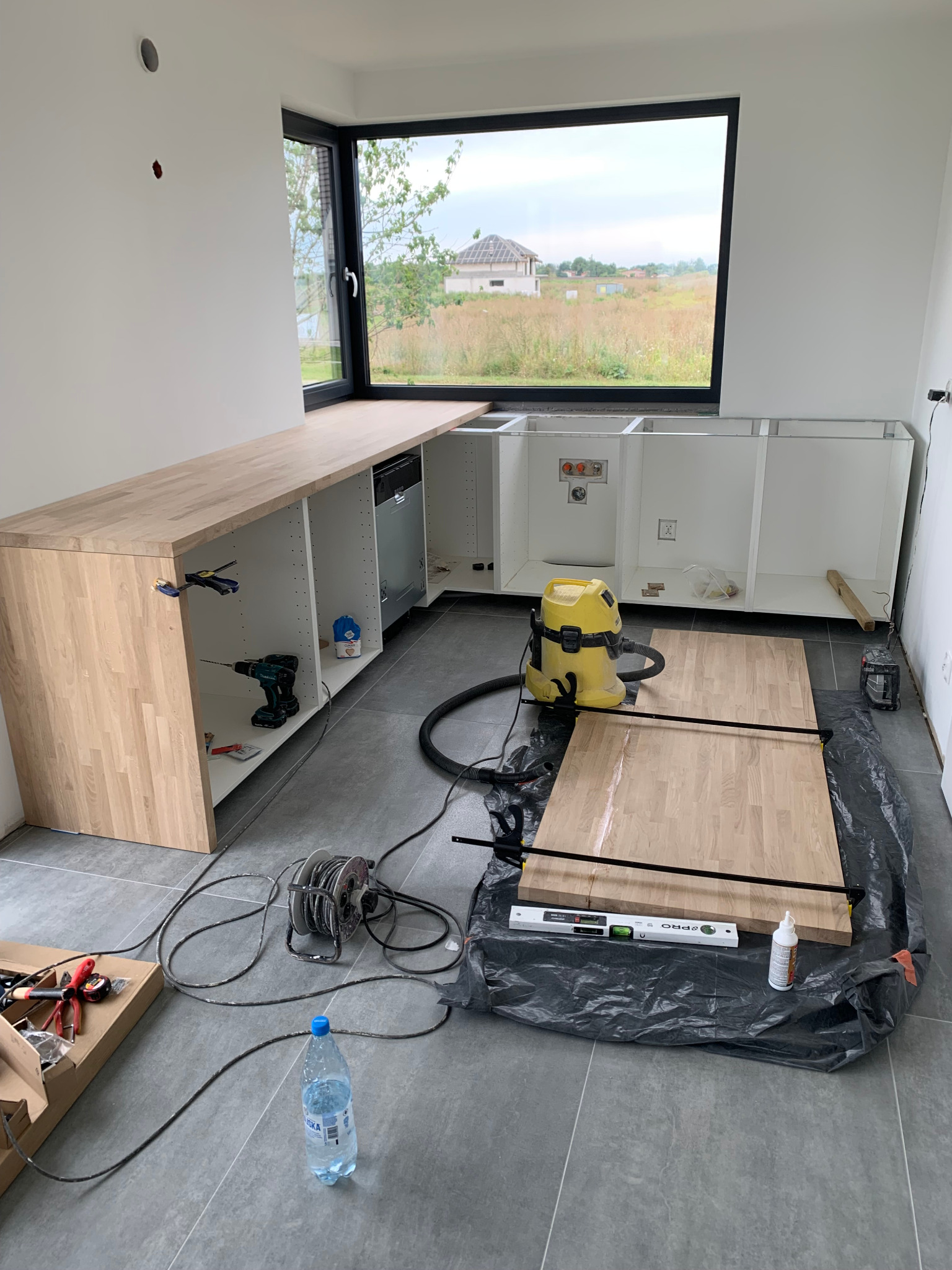 Montaż blatu kuchennego: widok na częściowo zamontowane szafki z drewnianym blatem, narzędzia i odkurzacz na podłodze z szarych płytek, okno z widokiem na krajobraz w tle.